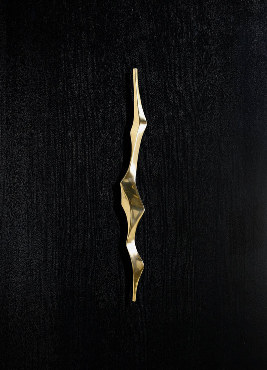A sculptural jewelry piece in polished gold-tone metal with an organic, undulating form, mounted on a black textured background.