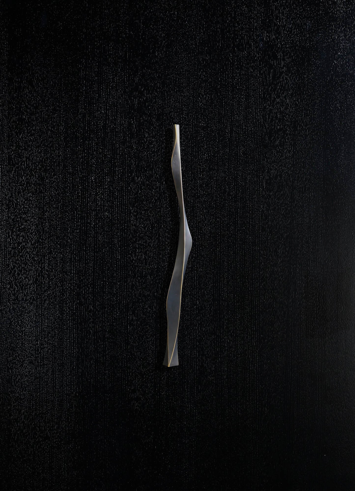 A sleek, modern sculptural object with a twisting, elongated shape in metallic silver finish, set against a textured black background.