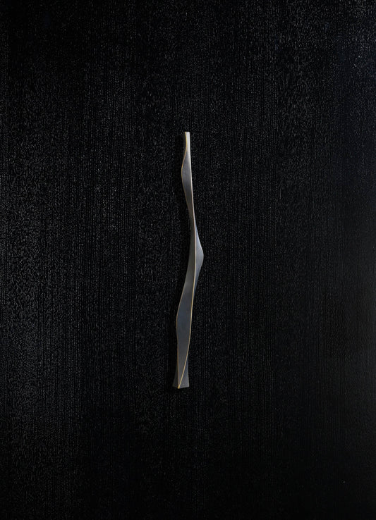 A sleek, modern sculptural object with a twisting, elongated shape in metallic silver finish, set against a textured black background.