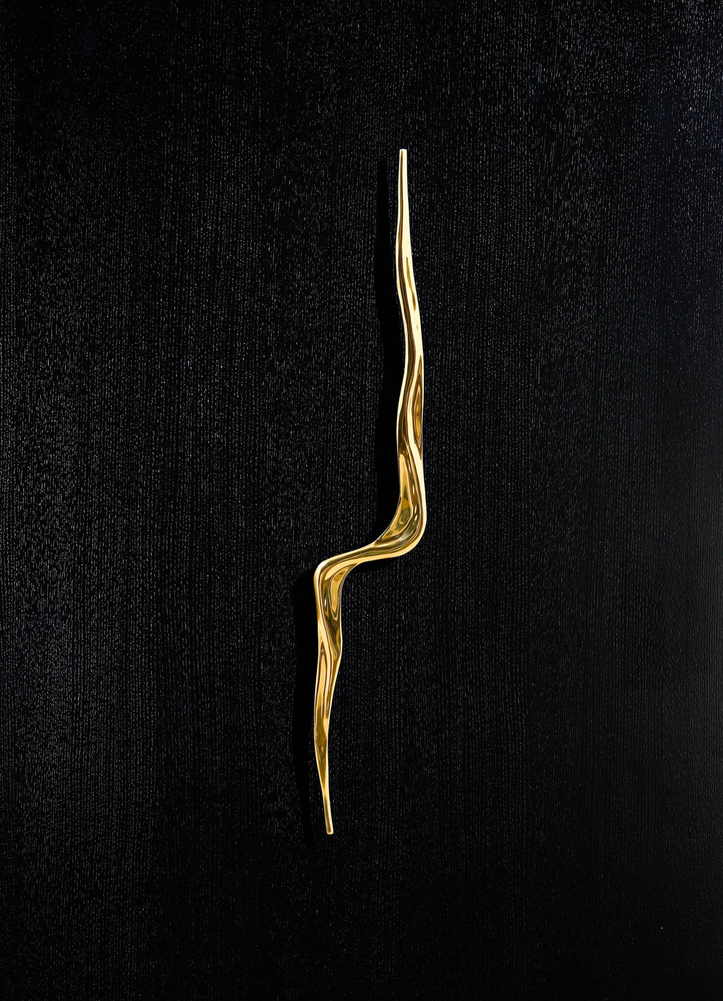 A sleek, gold-toned decorative sculpture with an organic, wavy form on a black textured background. The piece appears to be made of polished metal, emphasizing a contemporary abstract style.