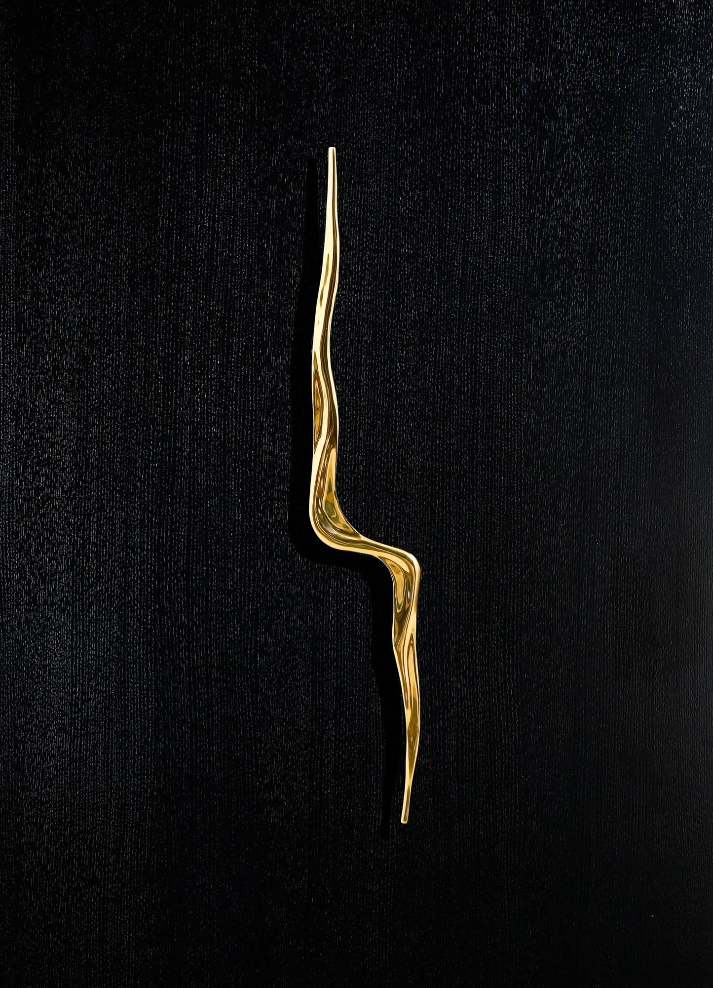 A gold-tone metal decorative item with an elongated, twisted, organic shape set against a black textured background.