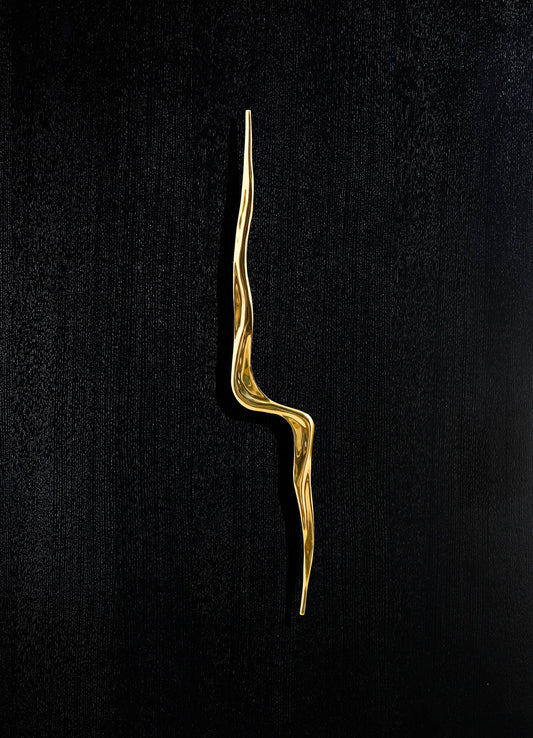 A gold-tone metal decorative item with an elongated, twisted, organic shape set against a black textured background.