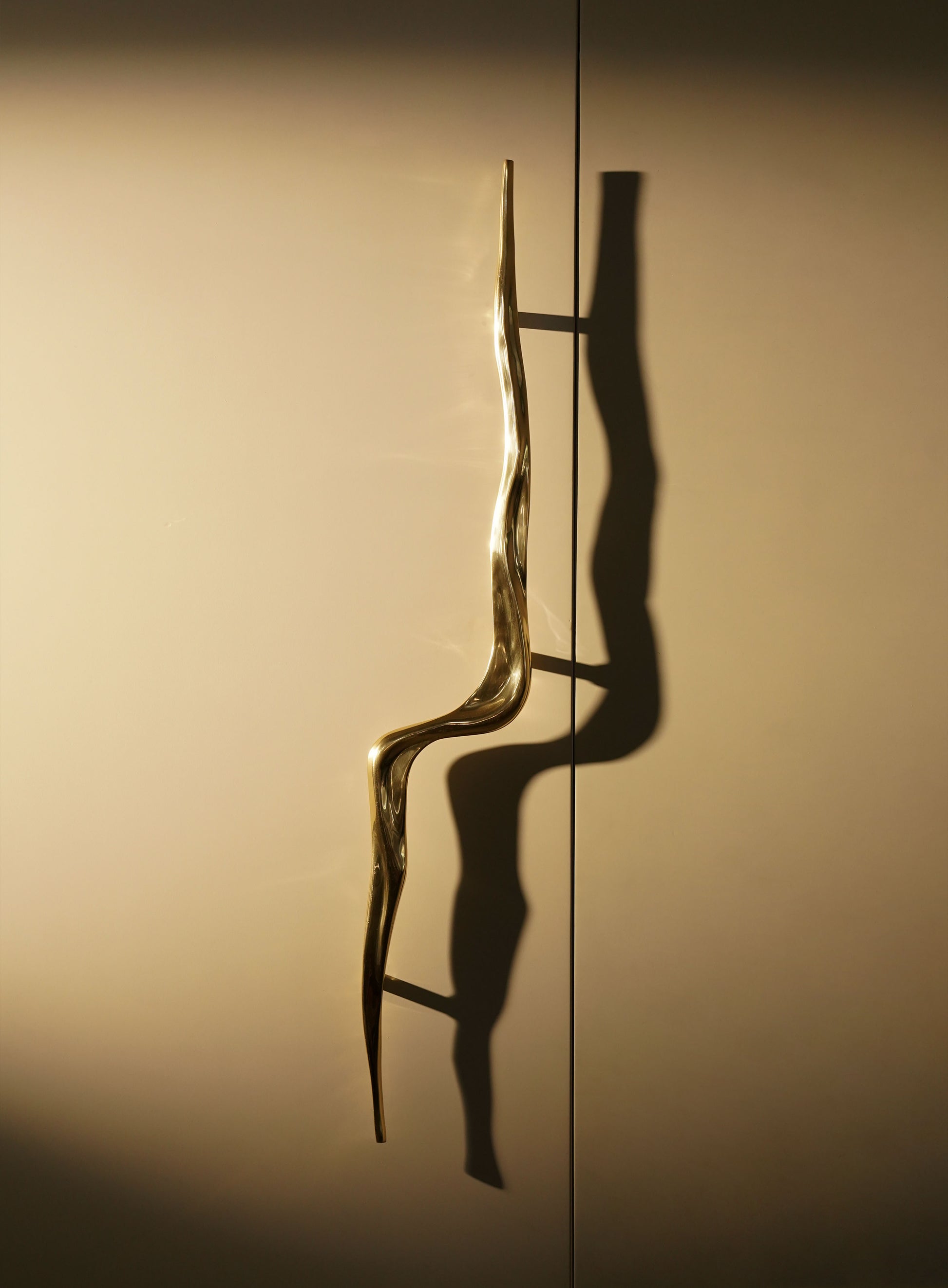 A decorative door handle with a wavy, sculptural design in a metallic finish, mounted on a smooth surface, creating elongated shadows.