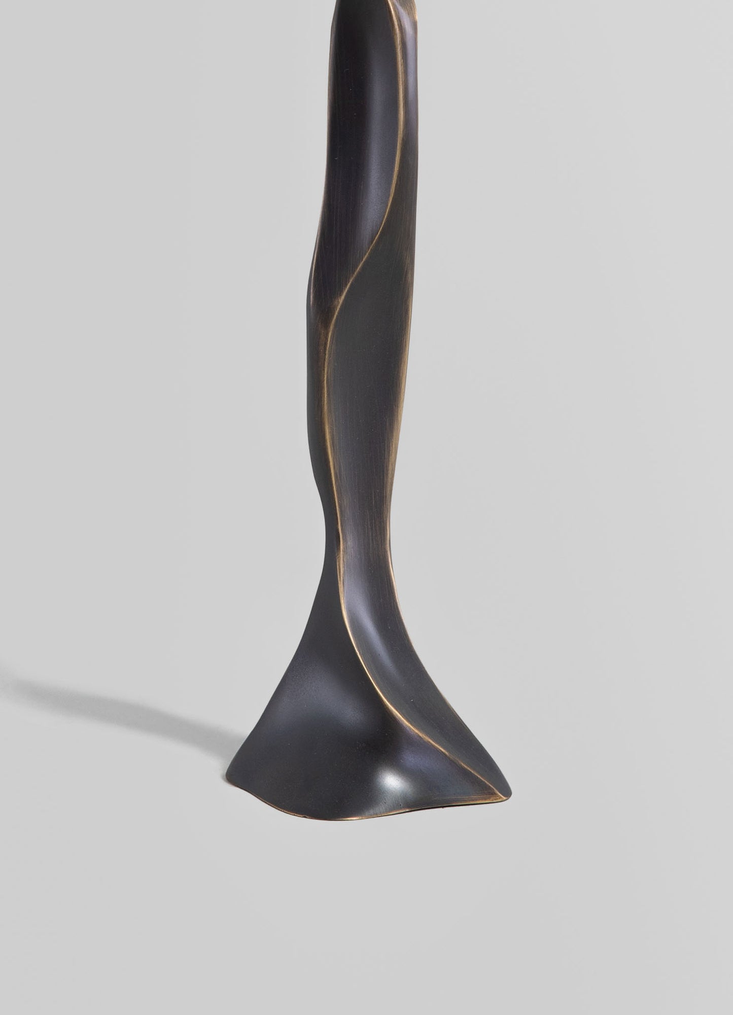 A sculptural table lamp with a twisted, elongated form made of dark finished wood or resin, featuring gold or brass accents along its curves, set against a neutral background. The style appears contemporary with a minimalist, artistic design focus.