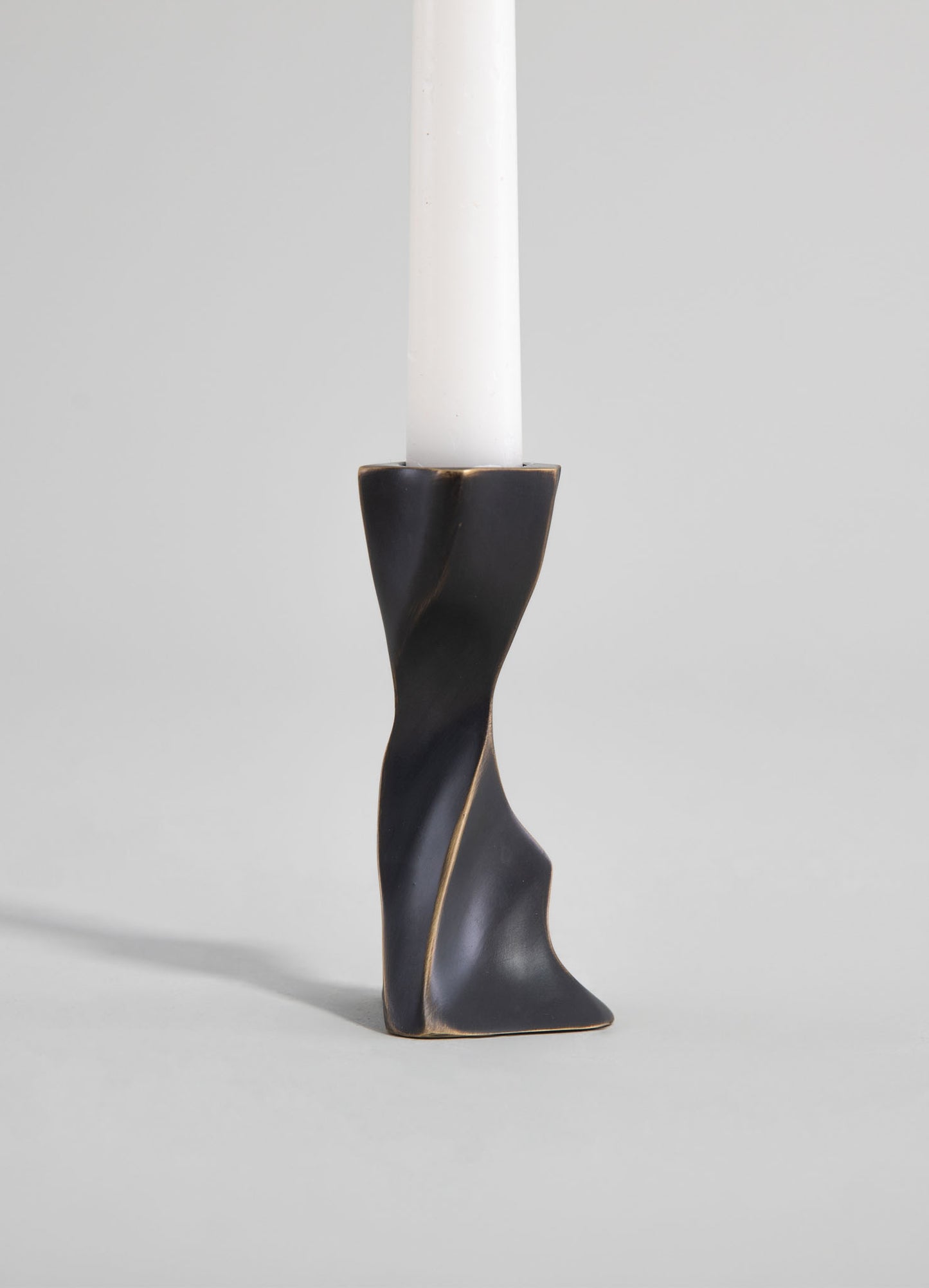 A sculptural candle holder with a twisted, abstract form in matte black and gold accents, designed to hold a tall white candle. The piece has a contemporary style with smooth, flowing lines.