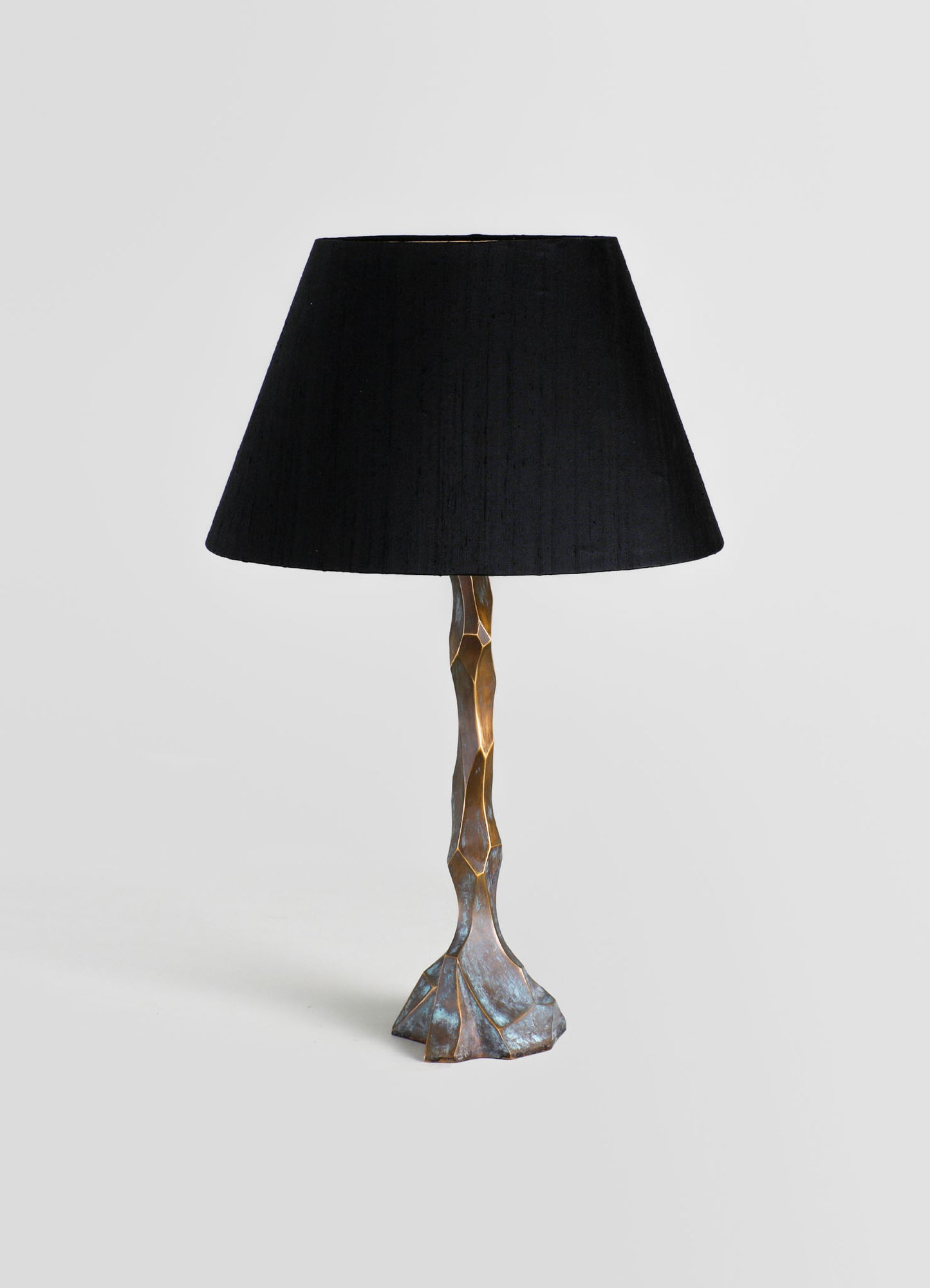 A table lamp featuring a sculptural metal base with a textured, geometric design and a black fabric lampshade, set against a plain background.