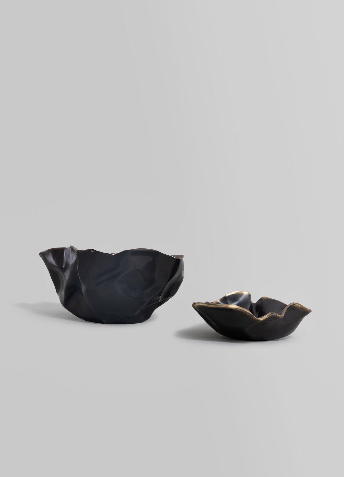 Two black ceramic bowls with irregular, organic shapes and a glossy finish, placed against a light gray background. The bowls have smooth, undulating edges and reflect light, emphasizing their sculptural design.