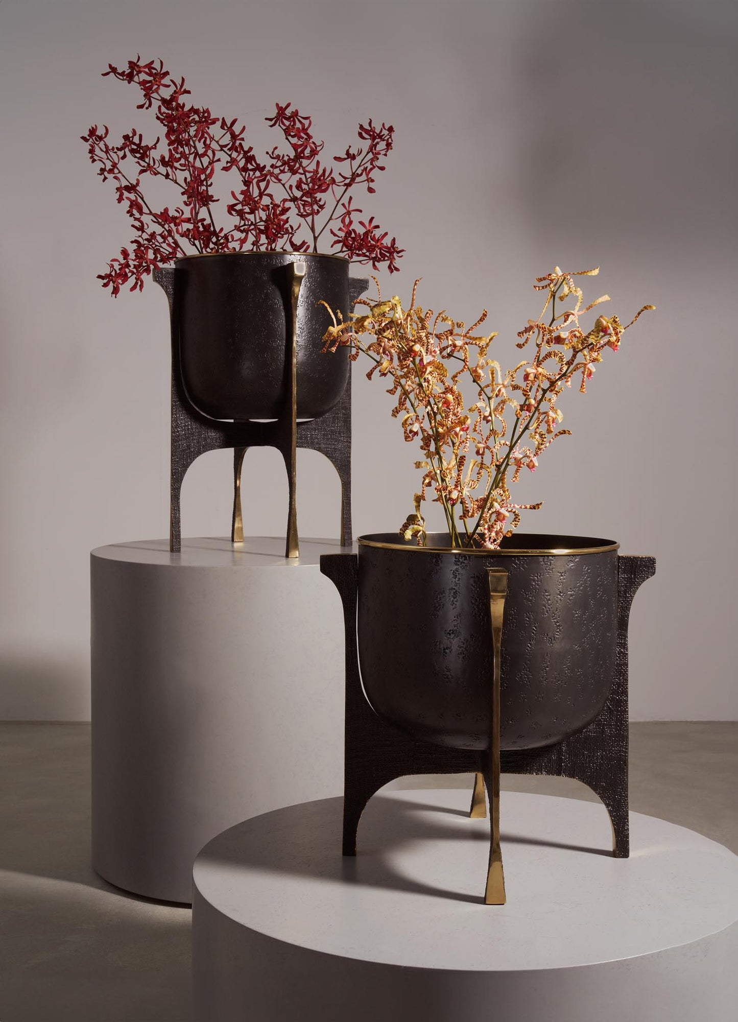 Decorative flower vases made of dark ceramic or metal with textured surfaces, featuring gold-tone accents on the supports and rims. The vases have a rounded, bowl-like shape with handles on the sides, displayed on white cylindrical platforms with contrasting textures.
