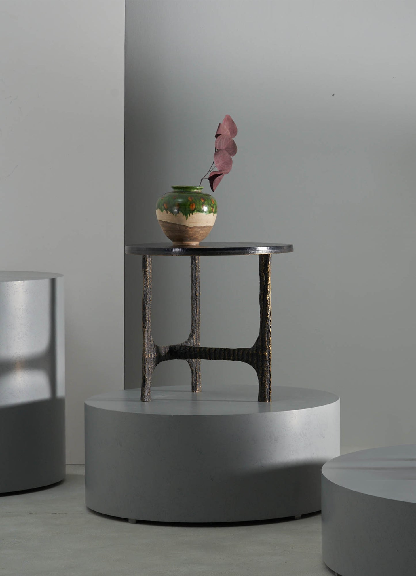 A ceramic vase with a rounded shape, glazed in a glossy green and beige finish with painted orange accents, holding dried pink leaves. The vase is placed on a black wooden side table with textured, curved legs, set against a neutral gray wall.