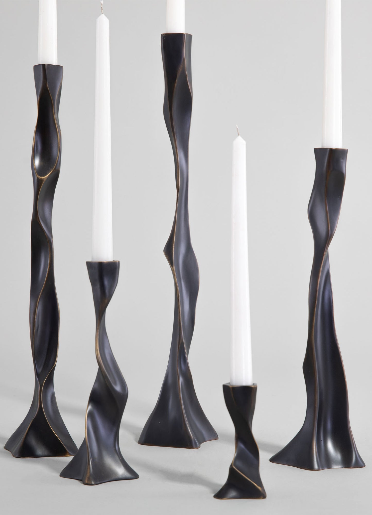 Sculptural candleholders made of dark metal with an undulating, twisted design, featuring a matte finish and holding tall white candles.