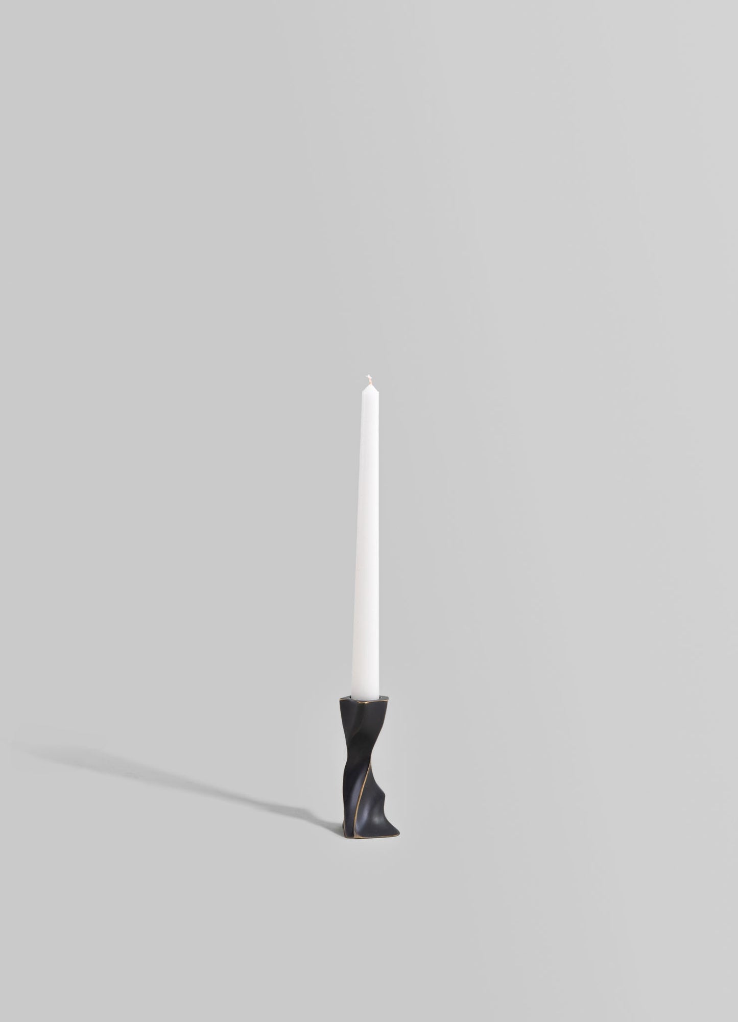 A modern candlestick holder with a sculptural black finish and gold accents, holding a white tapered candle, set against a light gray background.