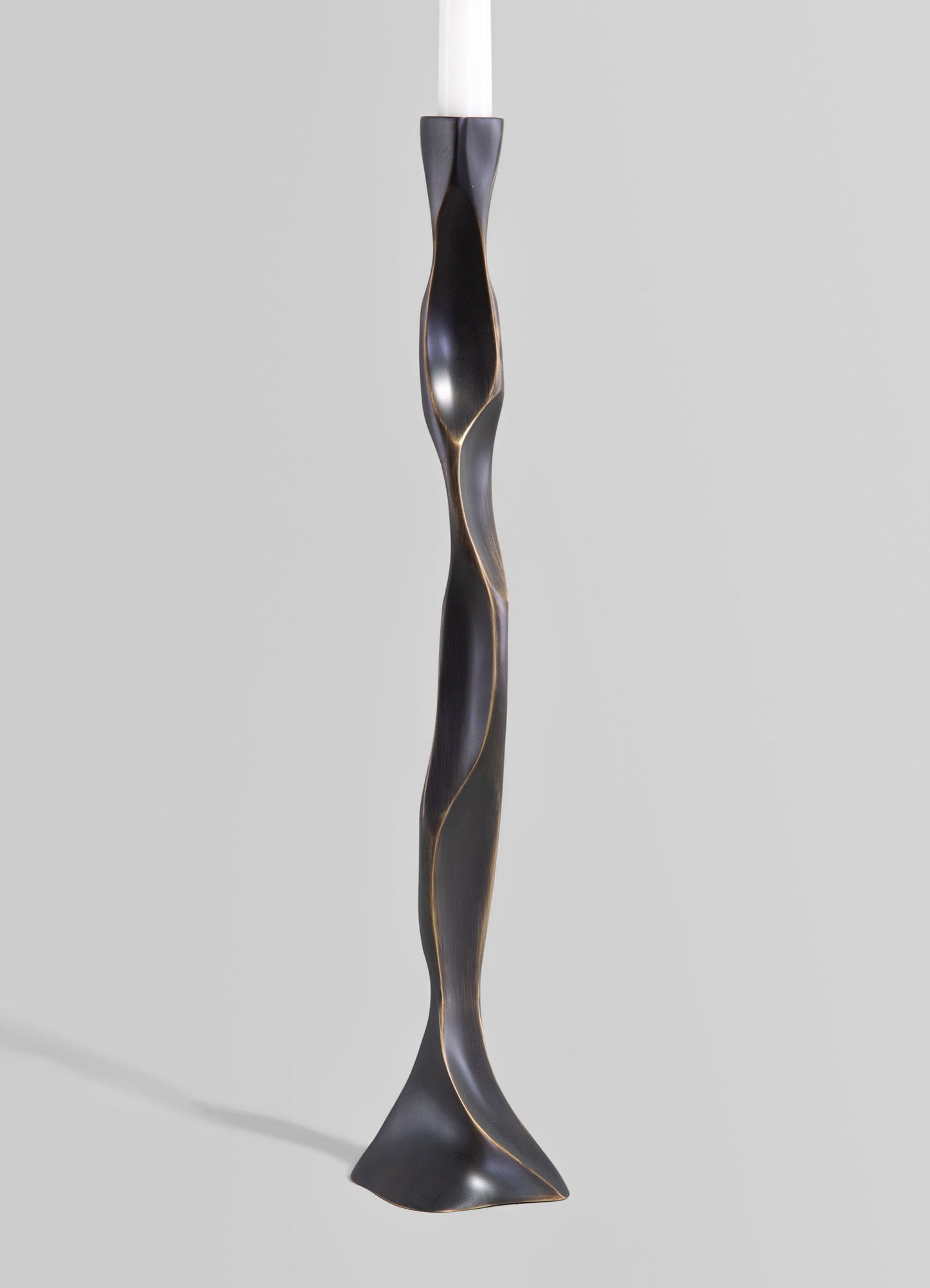 A sculptural candle holder with a tall, twisted form in dark metal finish, featuring subtle gold accents along the undulating curves, presented against a plain background.