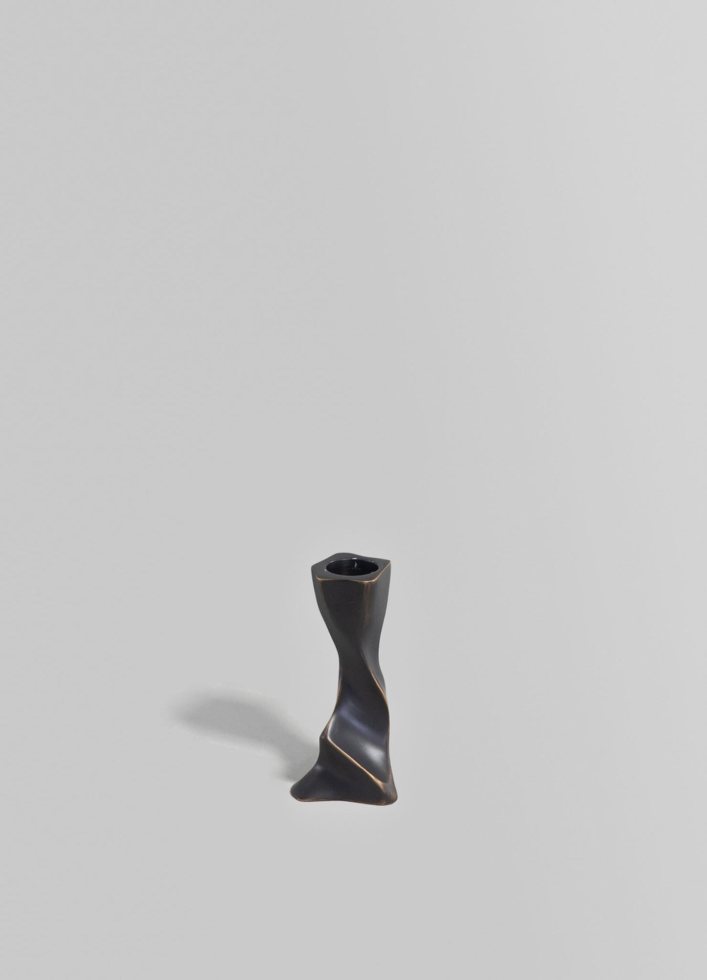 A black ceramic candlestick holder with a twisted, sculptural form, featuring a glossy finish and a square top for holding a candle, placed against a plain gray background.