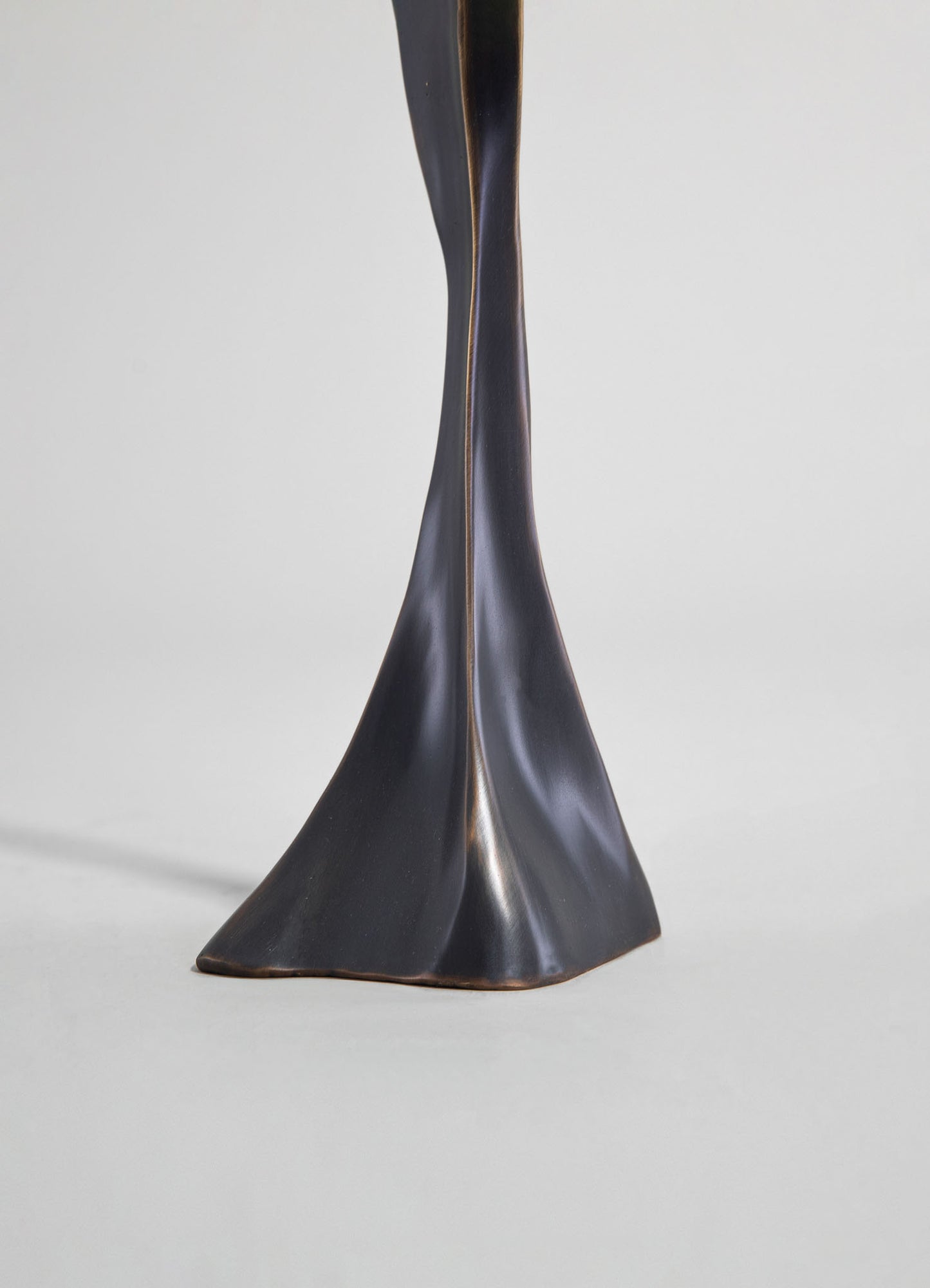 A close-up of a sculptural table or furniture leg made of dark-colored metal with a polished, curved design and subtle gold accents along the edges. The piece has an elegant, contemporary style with a sleek, artistic form.