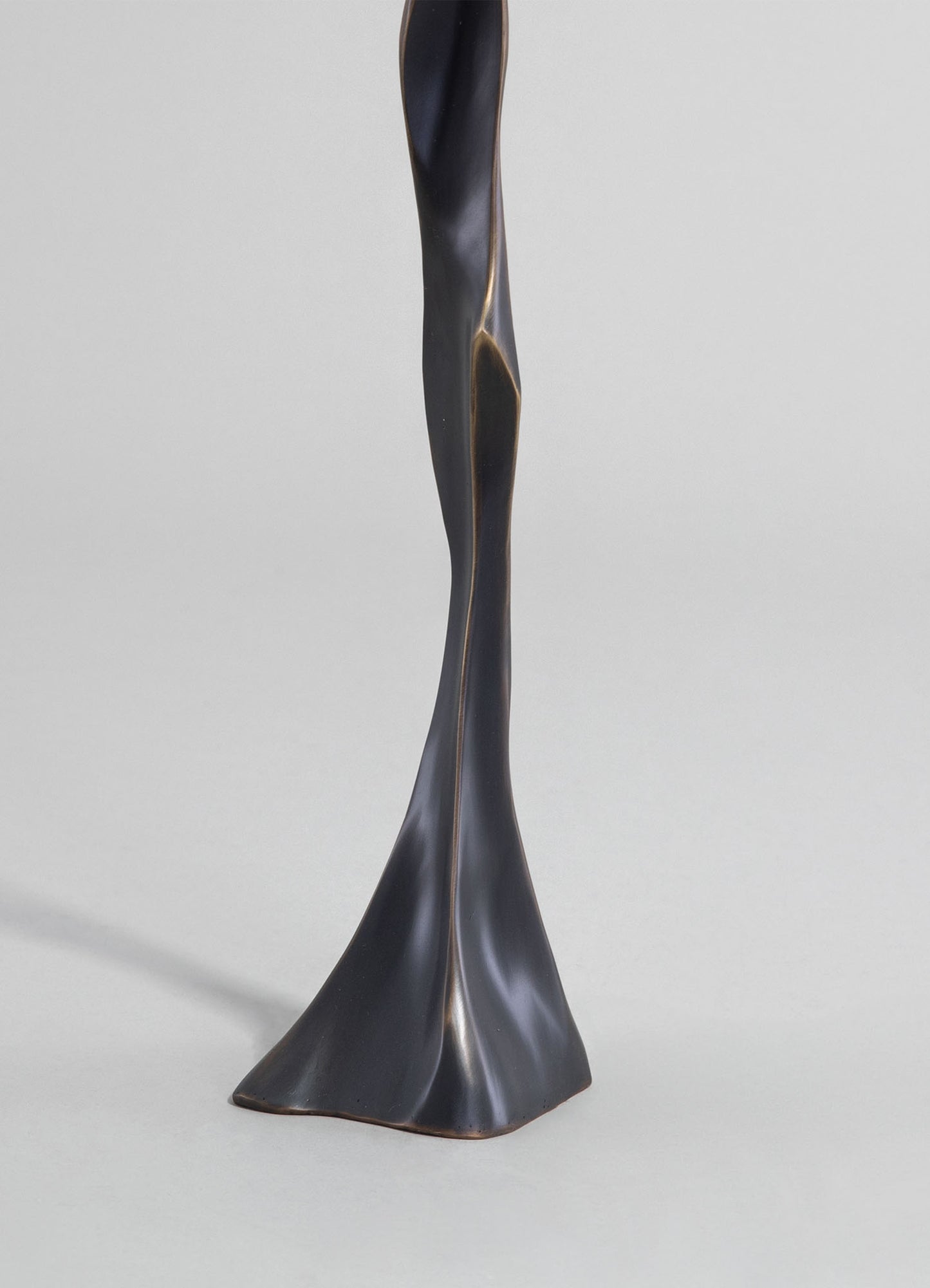 A sculptural table lamp with a twisted, organic form made of dark metal, featuring a smooth, matte finish. The design emphasizes contemporary minimalism and artistry, with an abstract, flowing silhouette.