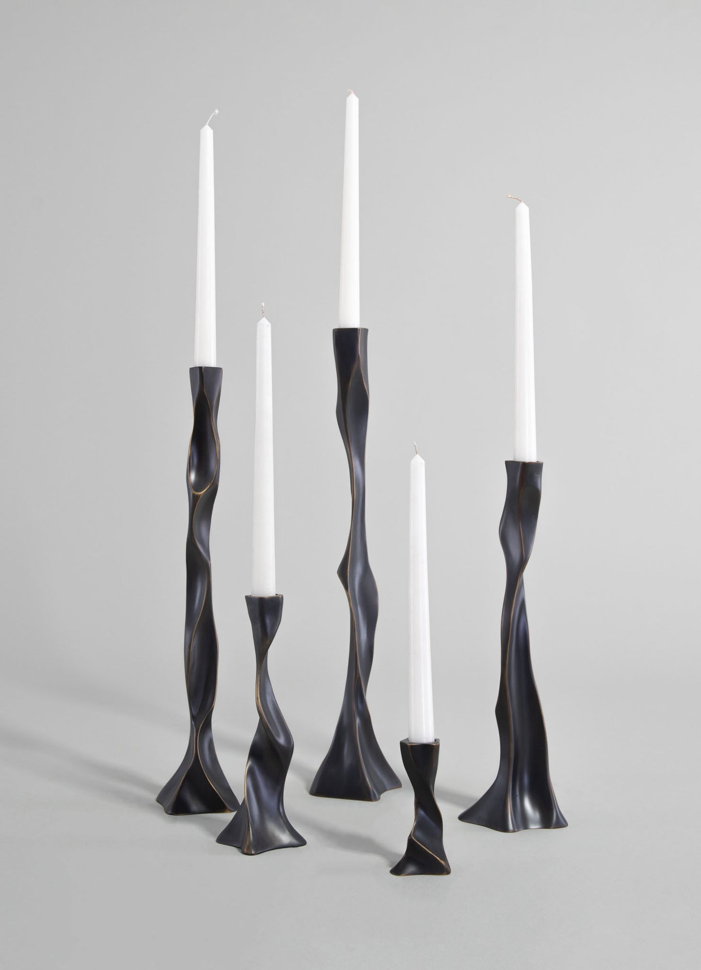 Modern black metal candle holders with twisted, sculptural shapes, each holding a tall white taper candle, arranged against a light gray background.