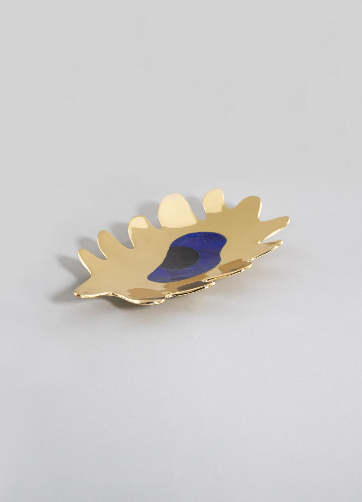 A decorative small dish made of gold-colored metal with a leaf shape and scalloped edges, featuring a blue and black stone or glass inset in the center.