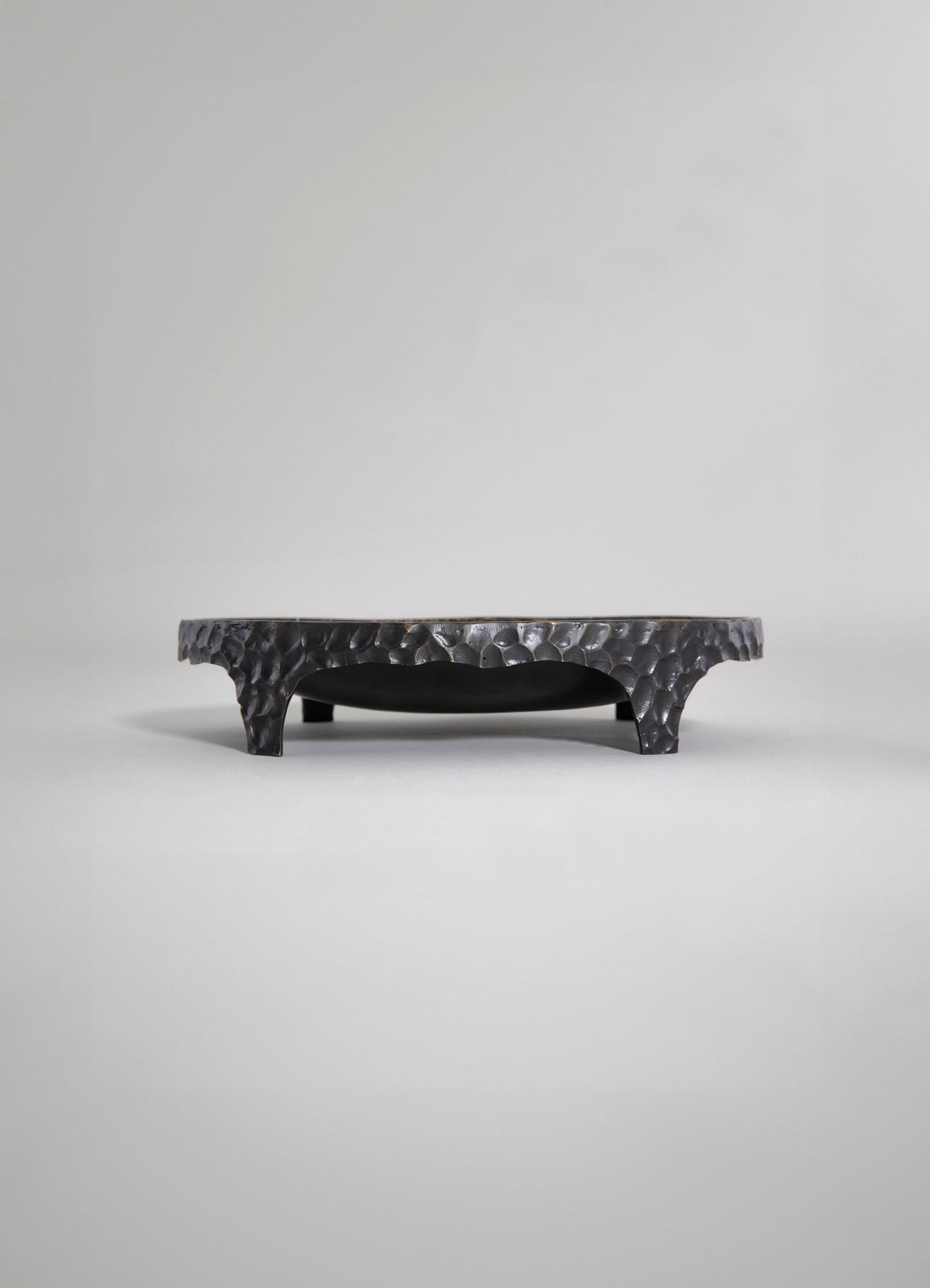 A black, textured tray with a hammered metal finish, featuring four curved legs and a smooth, slightly recessed interior surface, set against a plain light background.