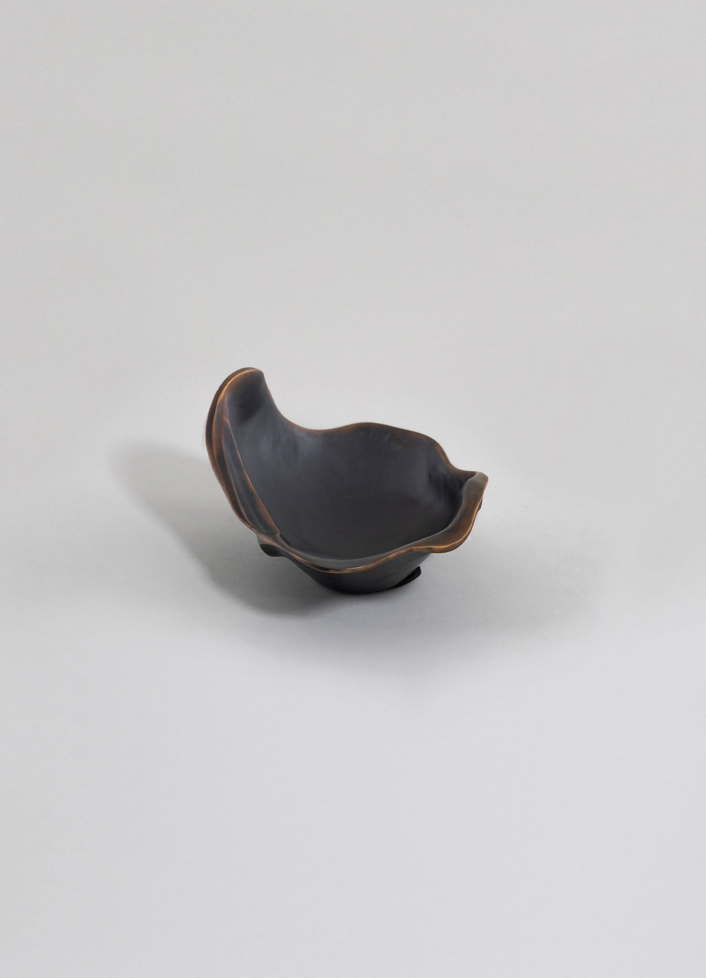 A black ceramic bowl with an irregular, organic shape and a subtle brown trim along the edges, set against a plain white background. The bowl has a matte finish and a minimalist, contemporary style.