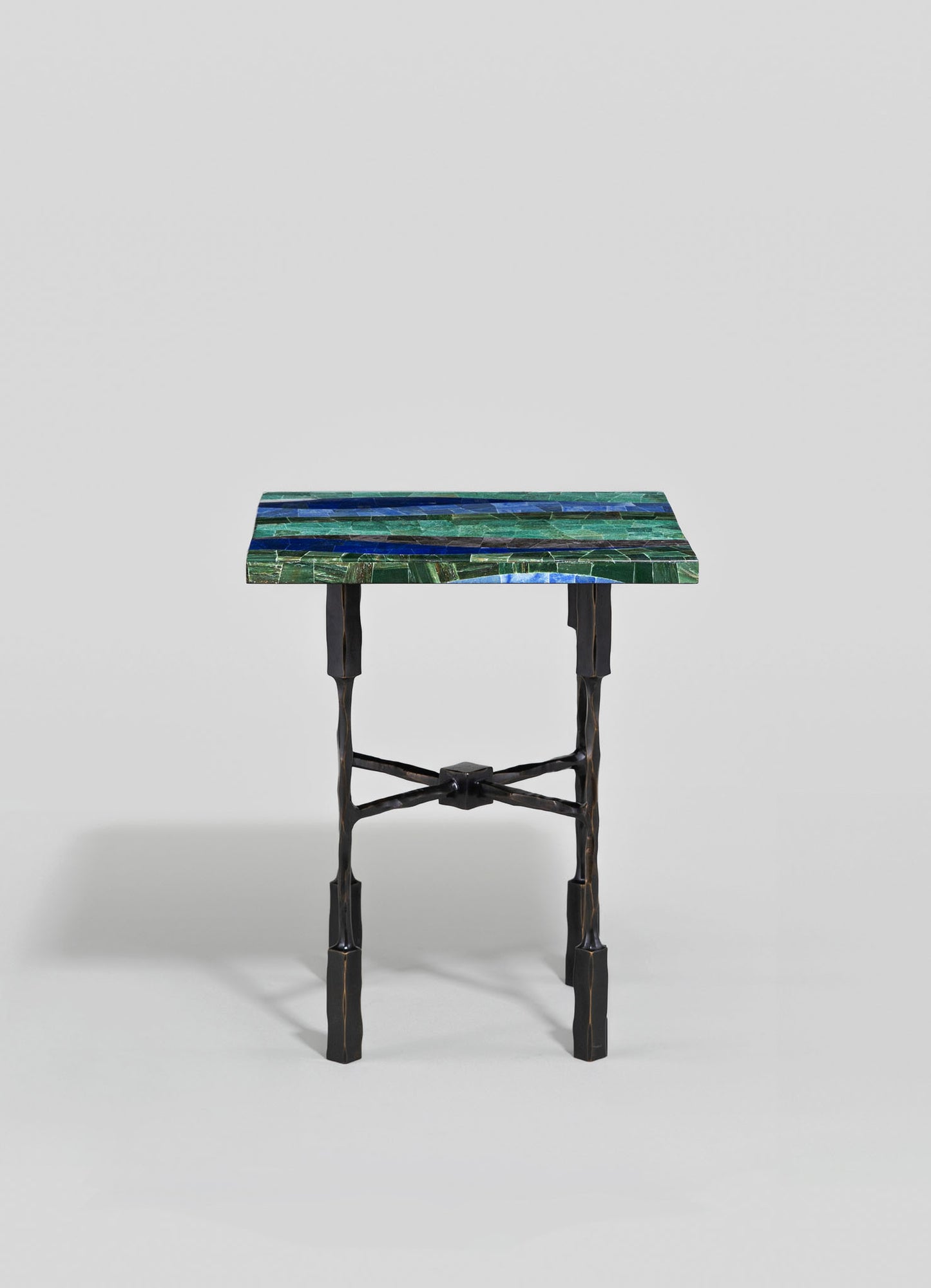 A small accent table with a rectangular mosaic top featuring shades of green and blue, supported by black, irregularly shaped wooden legs with cross-brace detailing, conveying a contemporary and handcrafted style.
