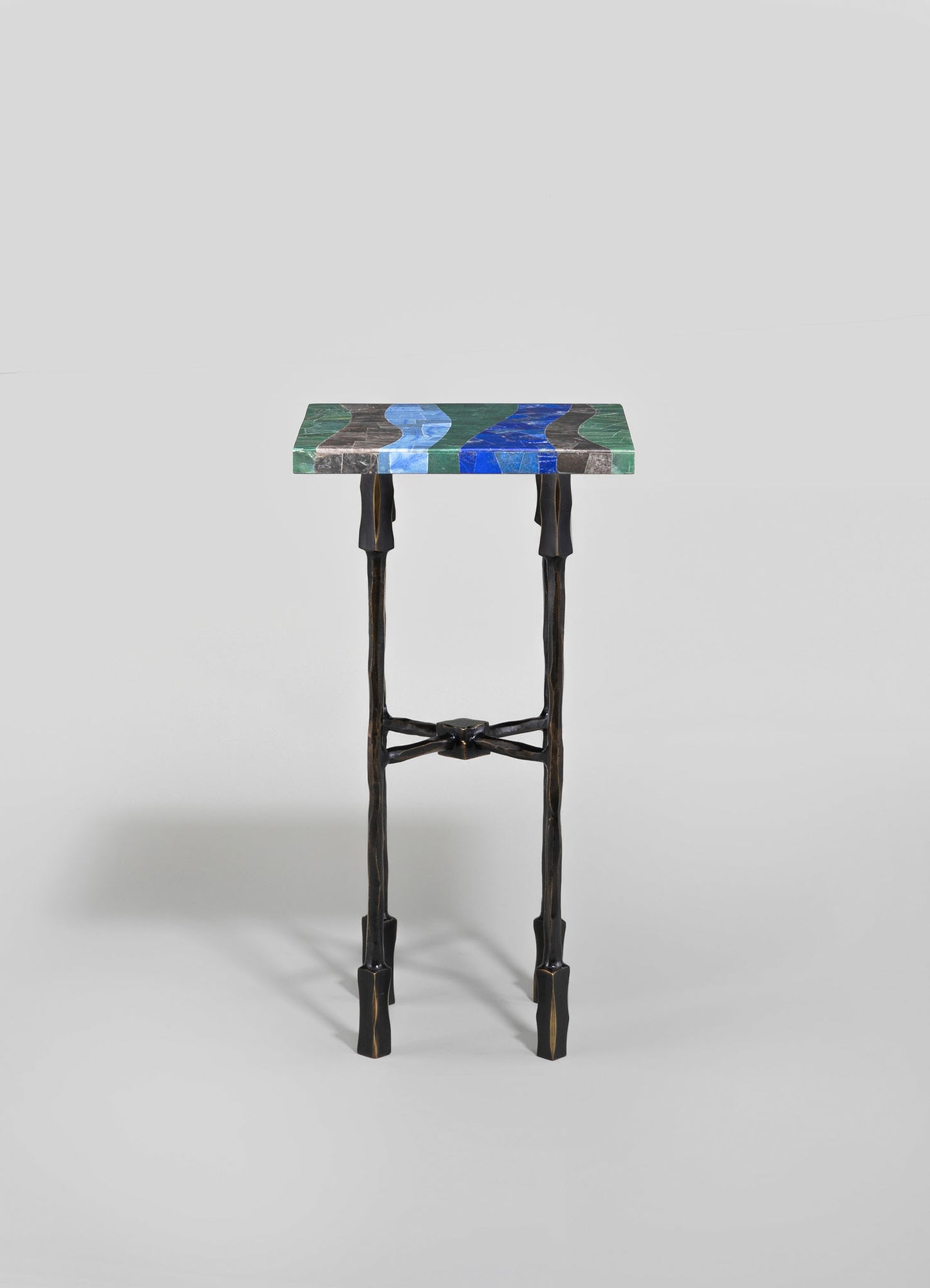 A small side table with a rectangular top made of multicolored stone in shades of blue, green, and gray, featuring organic, wave-like patterns. The table has dark, carved wooden legs with a slightly twisted design and cross supports, combining natural materials with an artisanal, contemporary style.