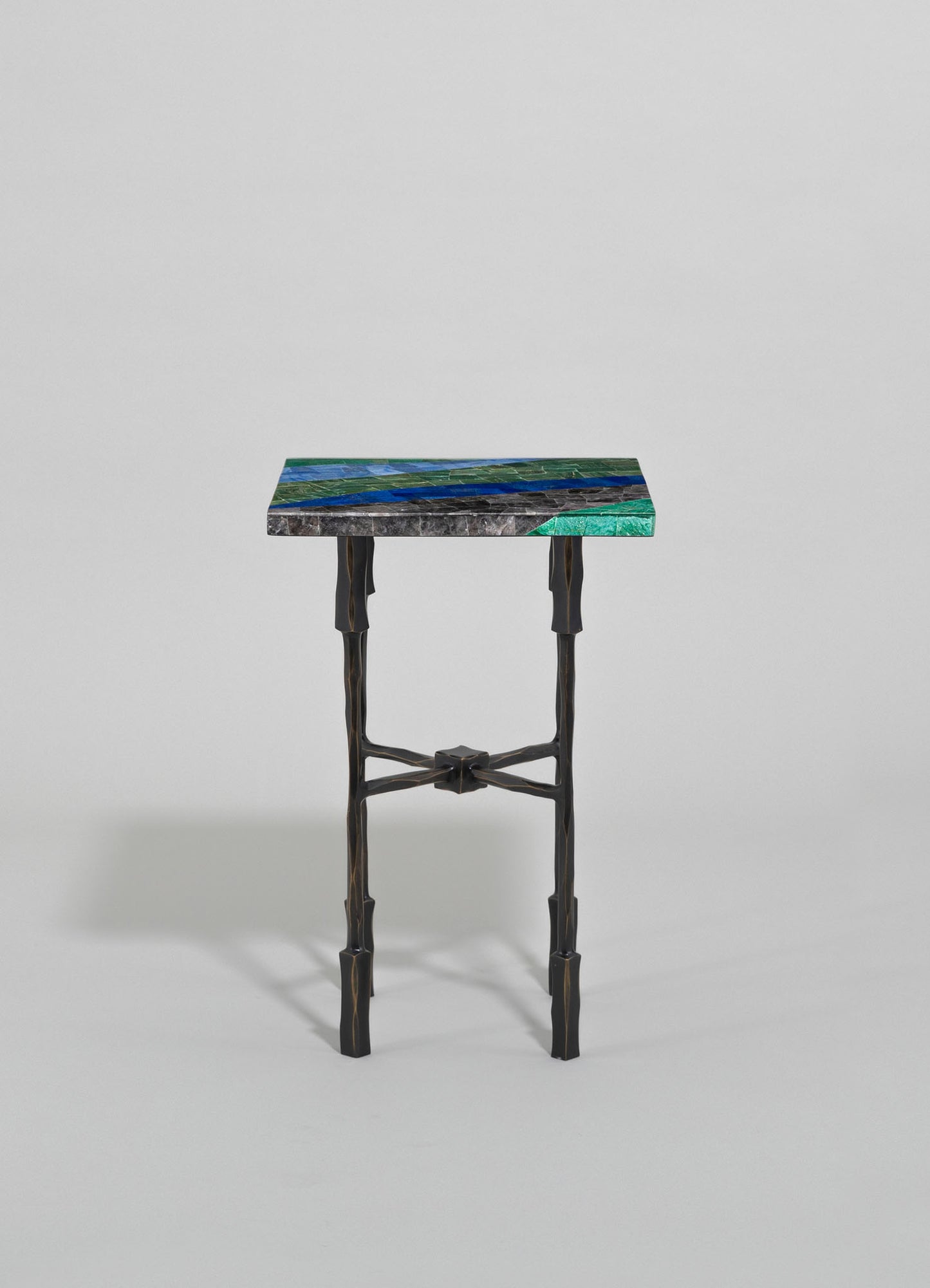A small side table with a rectangular top featuring a colorful, geometric mosaic pattern in shades of blue, green, and gray, supported by black, irregularly shaped wooden legs.