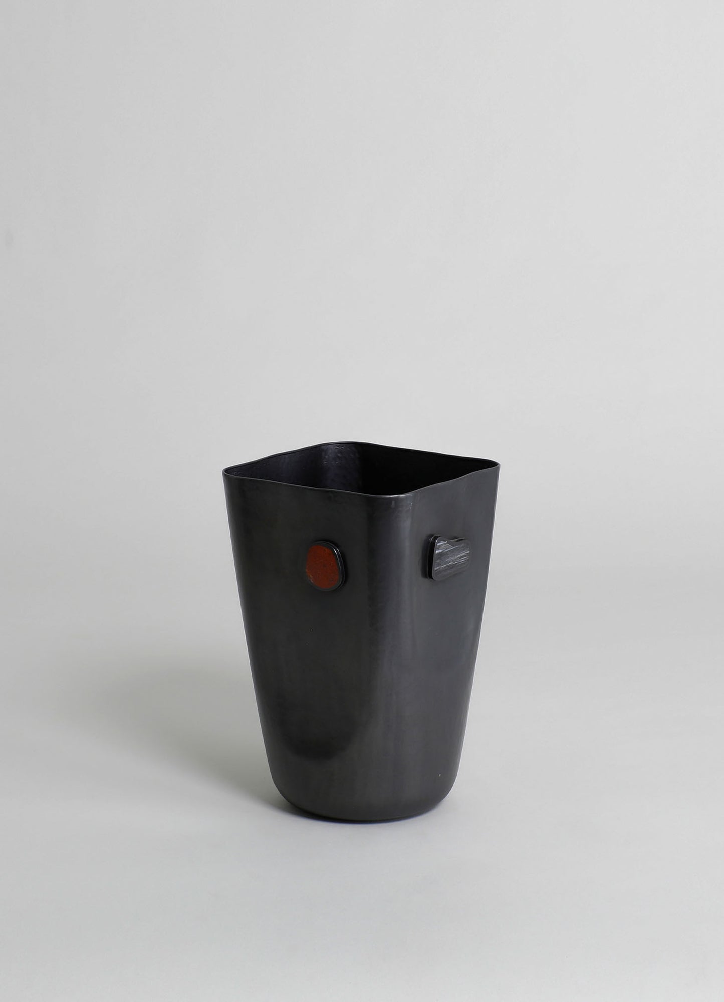 A black ceramic vase with a matte finish, featuring two small, rounded, colored accents on the side, set against a plain light background.