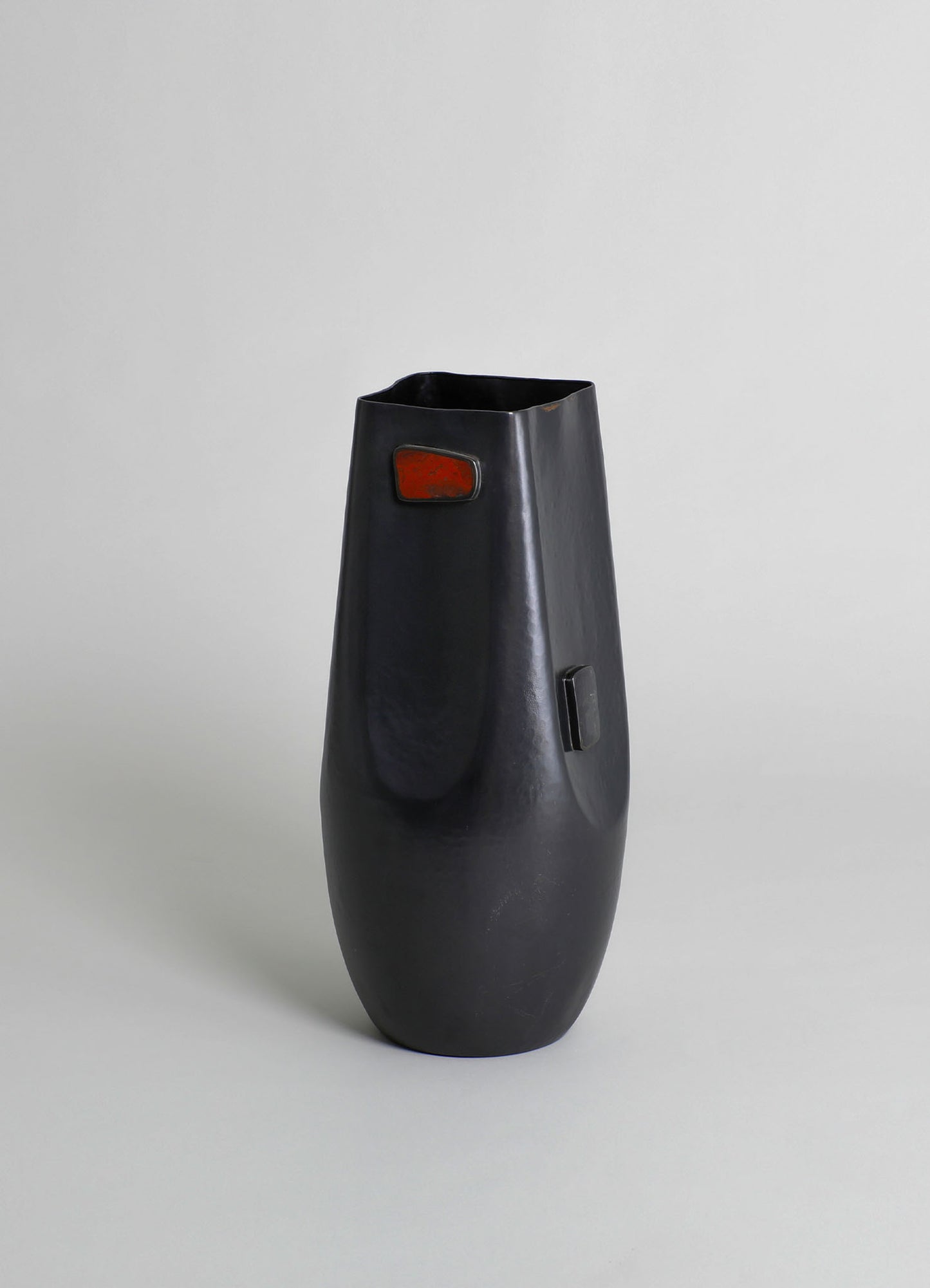 A black ceramic vase with a textured surface and a rounded, elongated shape, featuring a small rectangular orange patch near the top and a black handle or decorative element on the side.