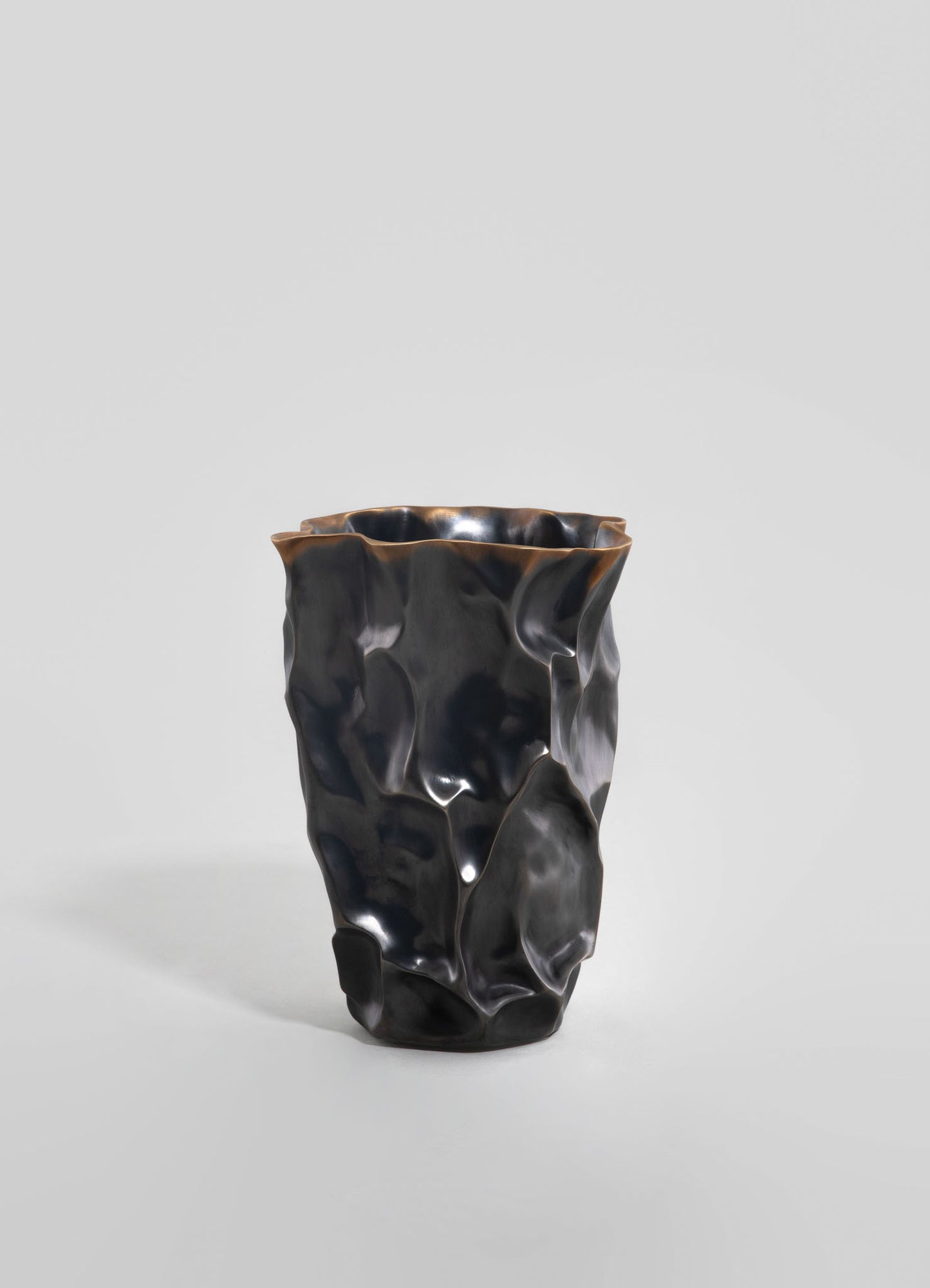 A sculptural vase made of dark, glossy ceramic or glass with an irregular, organic form and textured surface, featuring a wavy, uneven rim. The style is contemporary with an abstract, artistic motif against a plain background.