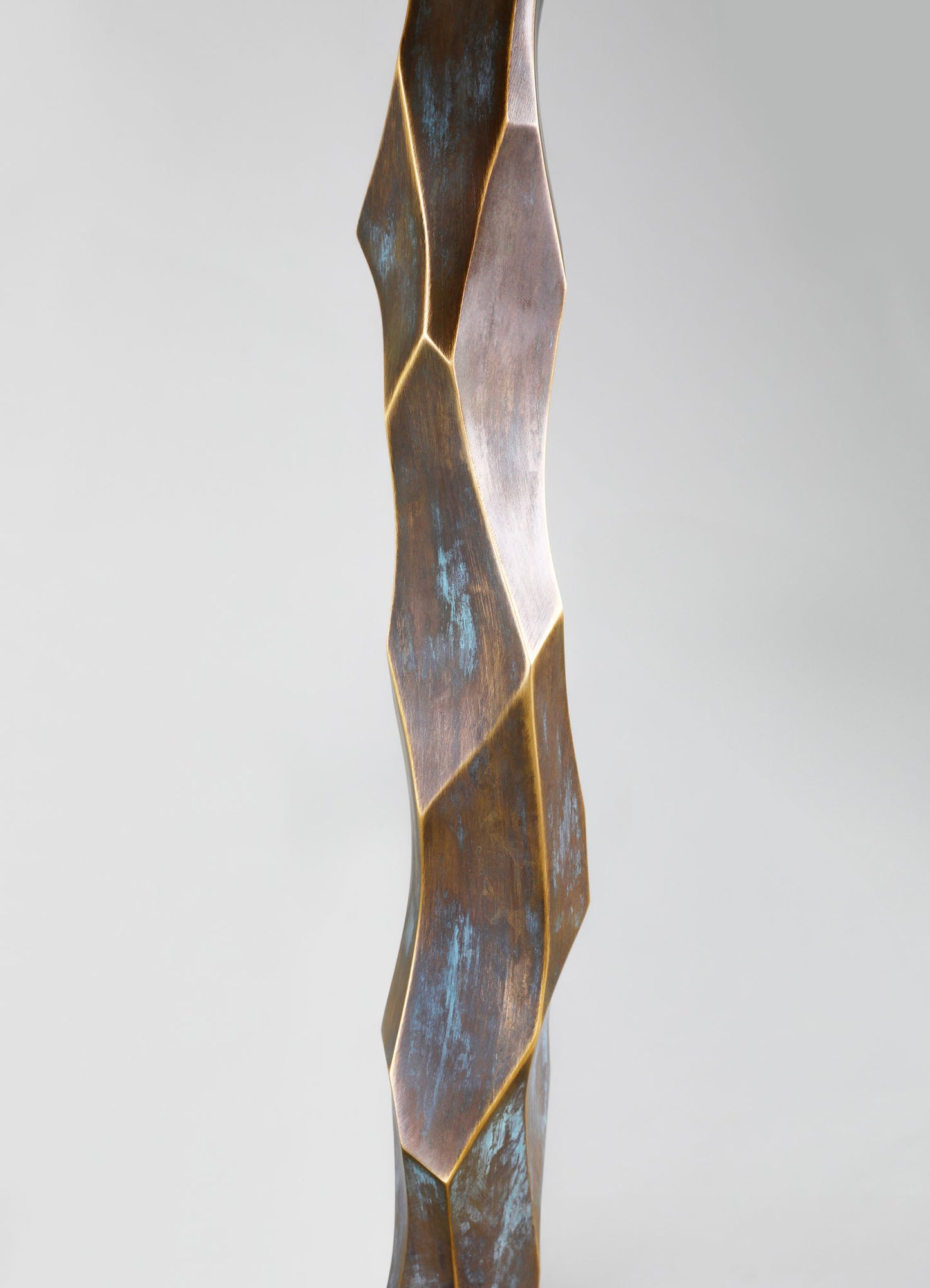 A sculpture made of metal with a twisting, faceted design featuring an oxidized, patinated surface with hints of bluish-green tones, and edged with gold highlights, in a contemporary artistic style.