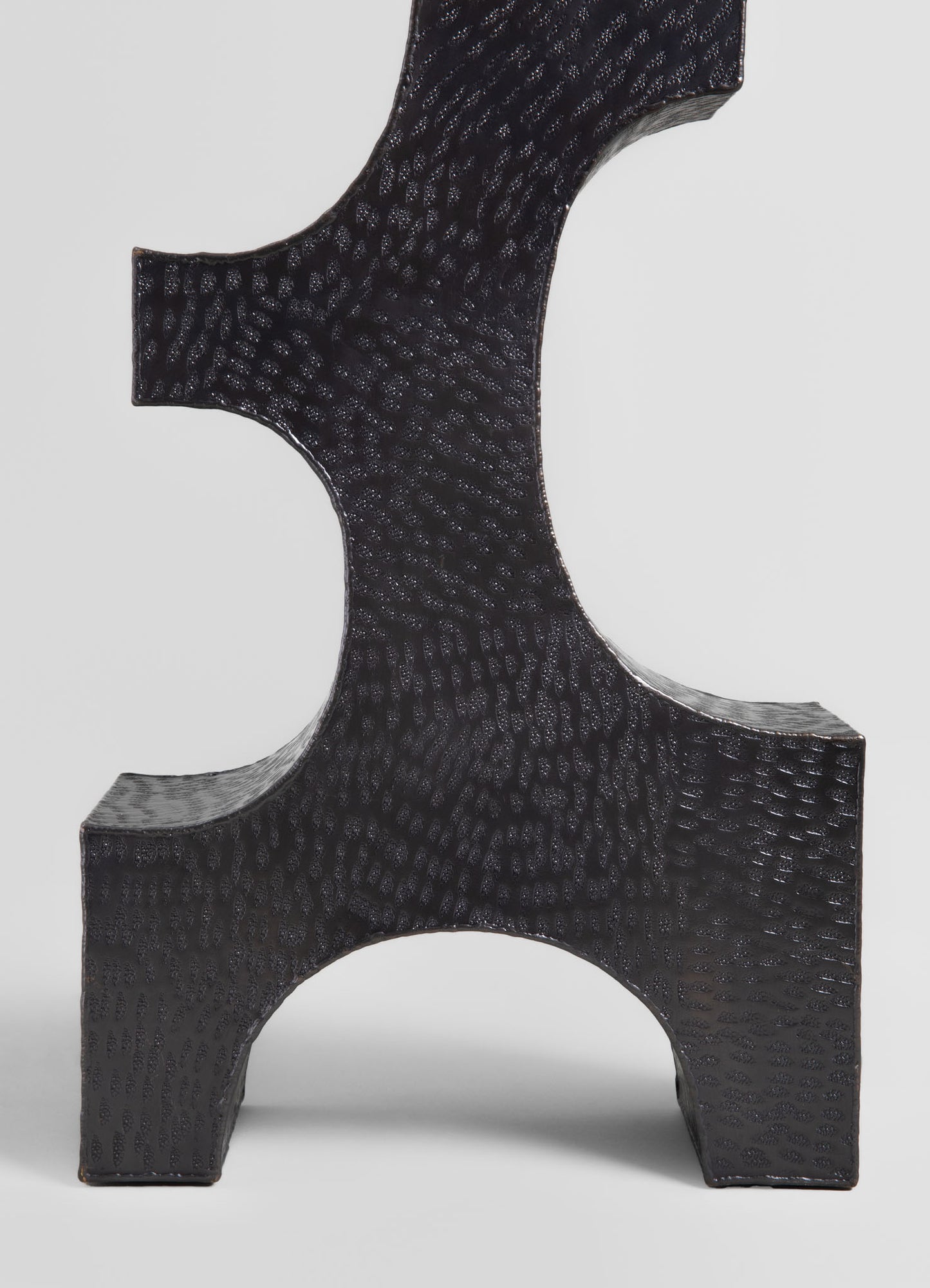 Black textured ceramic sculpture with a geometric, abstract form featuring curved and angular surfaces, set against a light background.