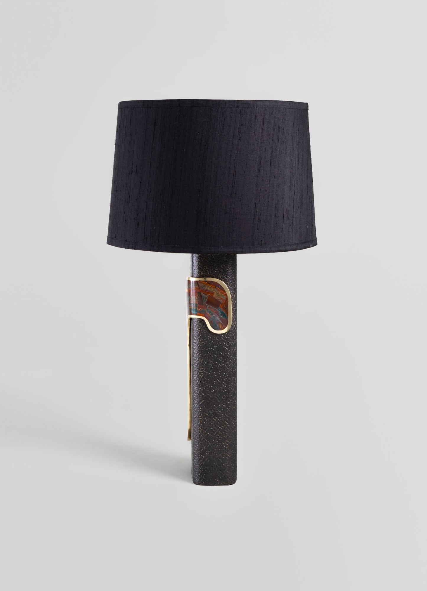A table lamp with a cylindrical black fabric shade and a textured dark metal base featuring an abstract decorative element in gold and multicolored accents.