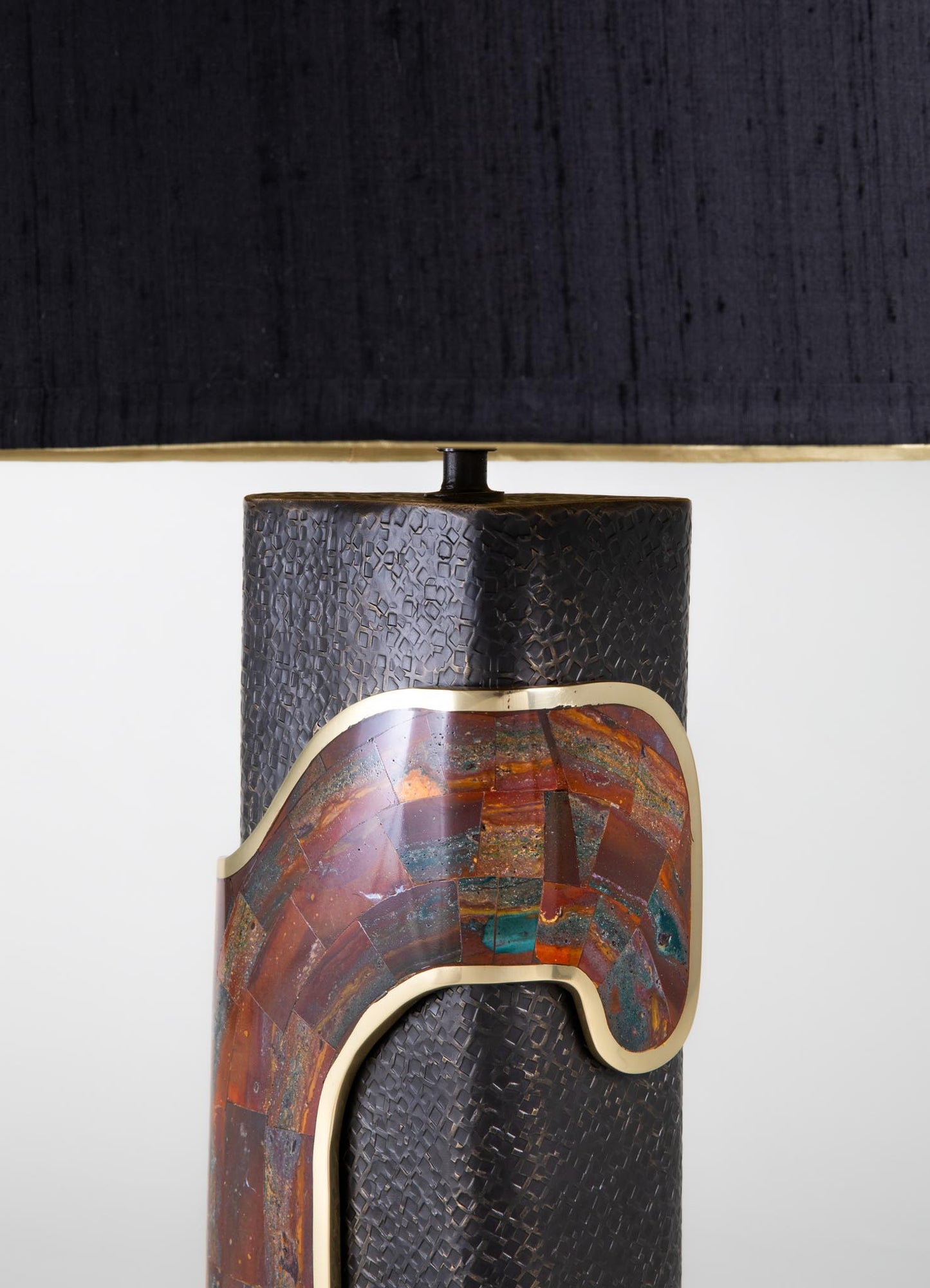 Decorative table lamp with a textured black ceramic base featuring a geometric pattern, accented by a multicolored stone inset with earthy tones, and topped with a black fabric lampshade.
