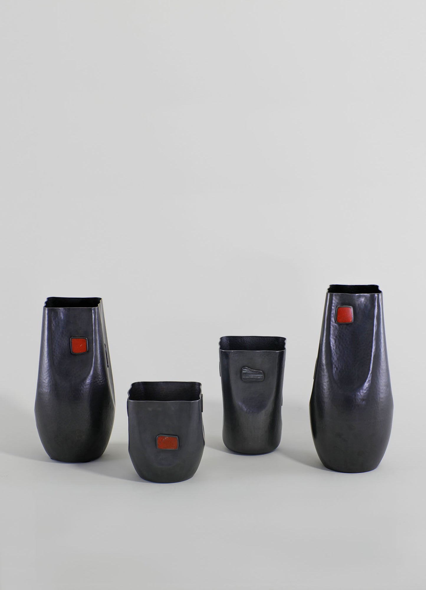 Set of four black ceramic vases with textured surfaces and rectangular reddish-orange accent tiles, featuring a modern minimalist style. The vases vary in height and shape, with some having a rounded form and others being more rectangular.