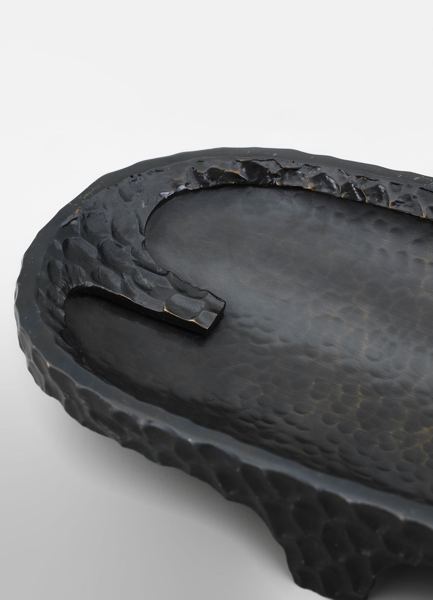 A black, textured ceramic tray with a raised, irregular rim and a smooth central surface. The tray features a hammered pattern on the border and a matte finish, conveying a handcrafted and rustic style.