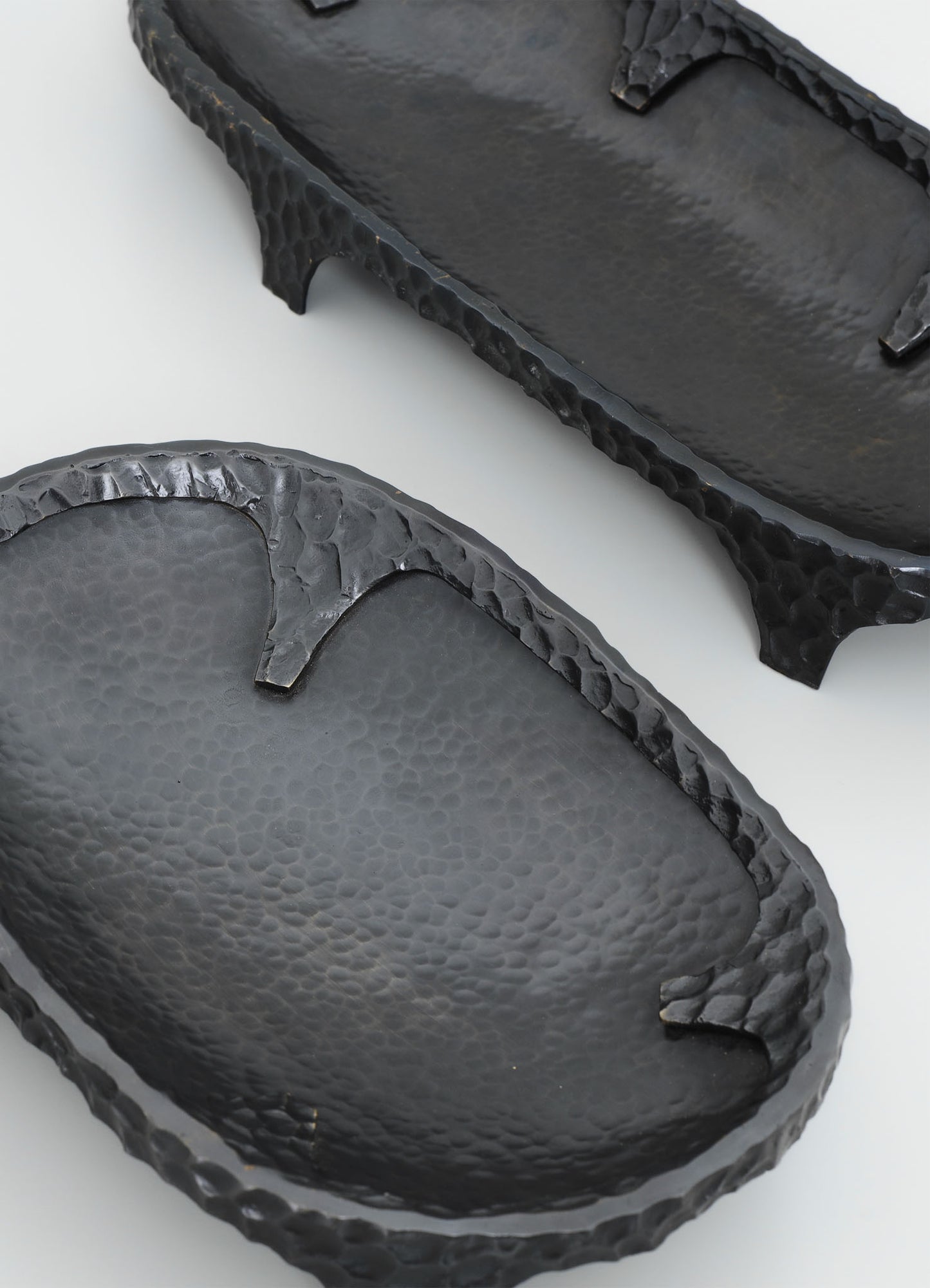 Black hammered metal trays with textured surfaces and scalloped edges, one oval and one rectangular, on a white background.