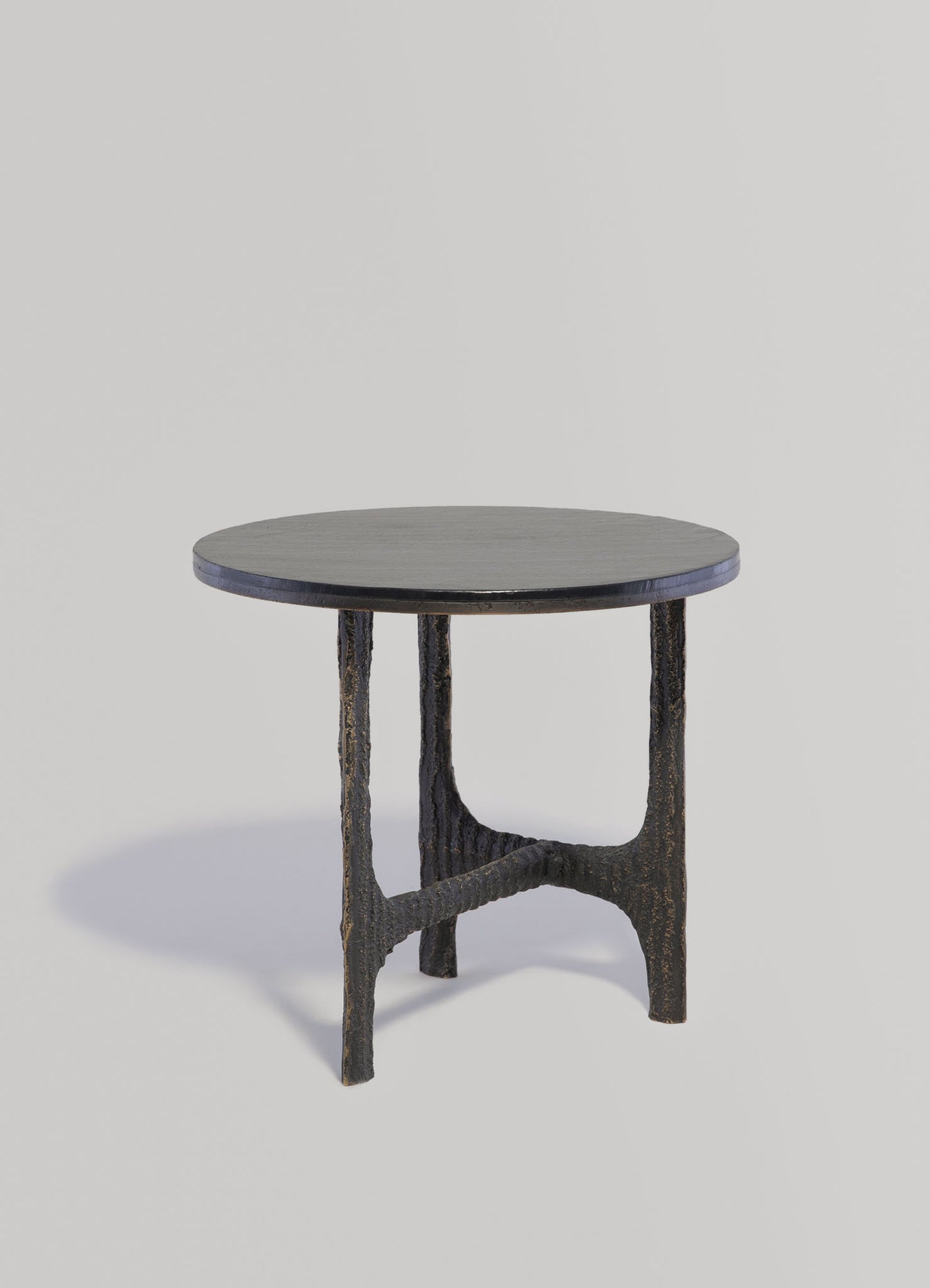 A small round table with a dark, textured wood top and rustic black metal legs featuring a rough, distressed finish. The design combines minimalist shape with a vintage, handcrafted style.