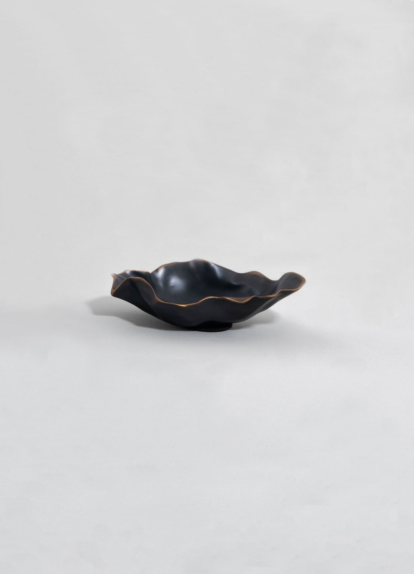 A black ceramic bowl with a wavy, irregular edge and a subtle metallic finish, set against a plain background. The bowl has an organic shape and a smooth, glossy surface.