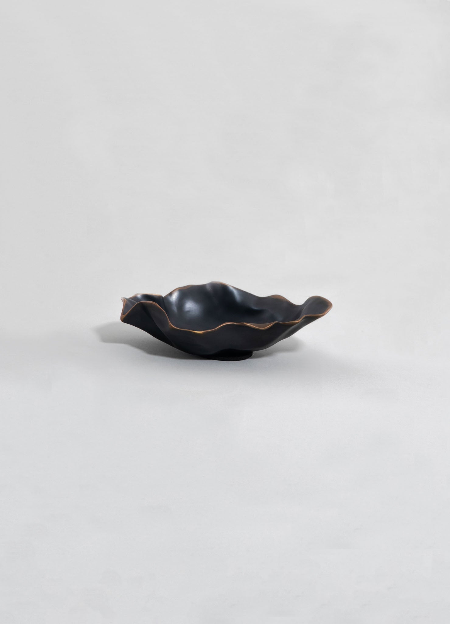 A black ceramic bowl with a wavy, irregular edge and a subtle metallic finish, set against a plain background. The bowl has an organic shape and a smooth, glossy surface.