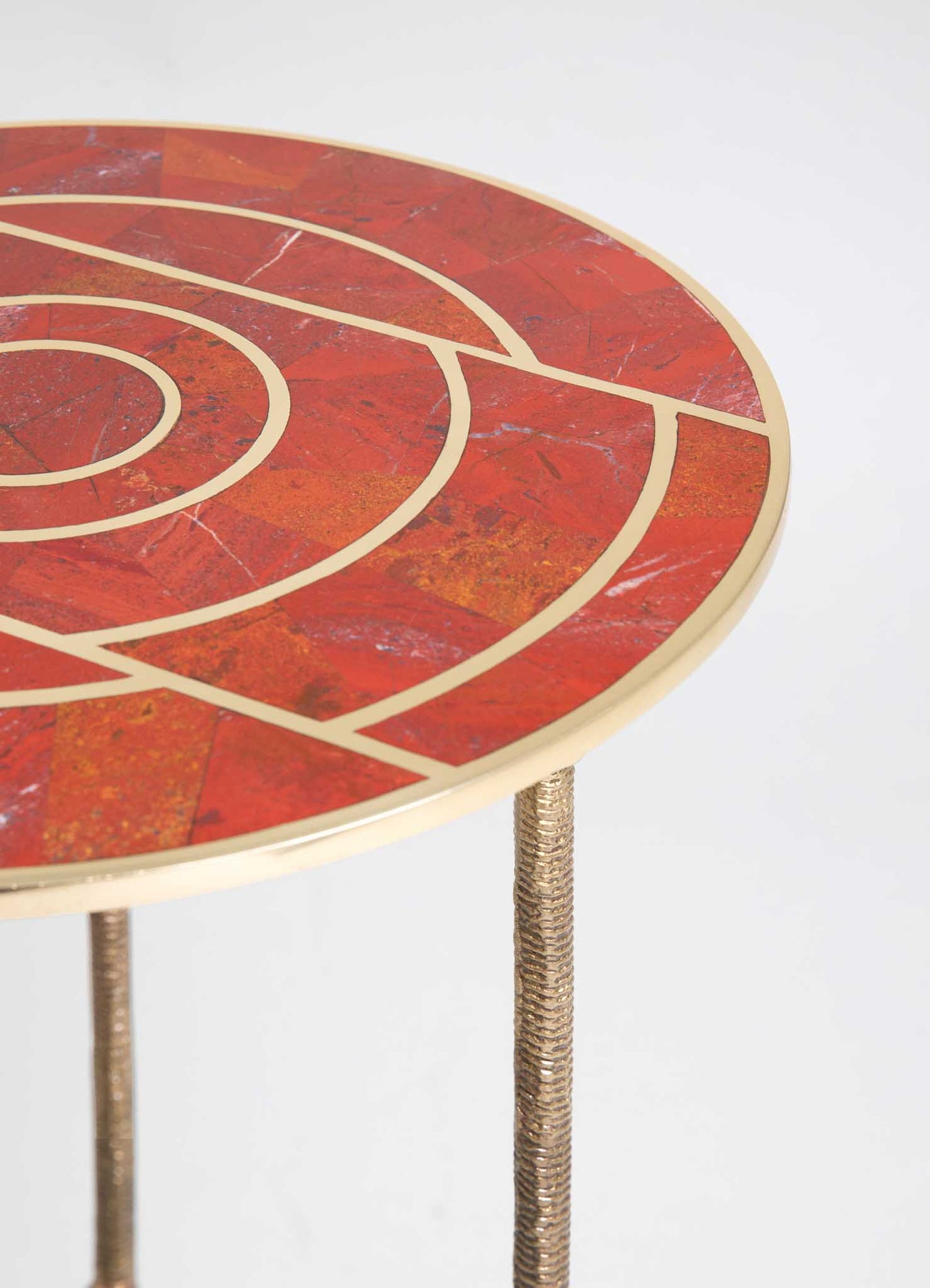 Round side table with a red marble top featuring beige geometric inlay patterns and a gold trim. The table has three textured metal legs with a bronze finish.