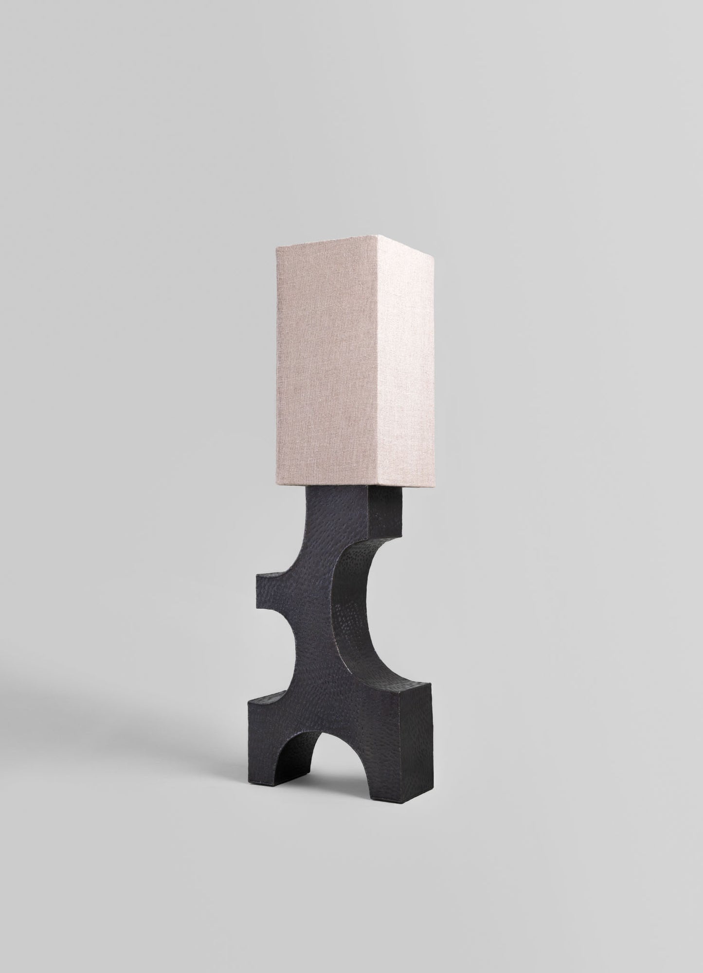 A table lamp with a block-shaped beige fabric shade and a curved black wooden base, featuring a modern design with smooth lines and contrasting neutral tones.