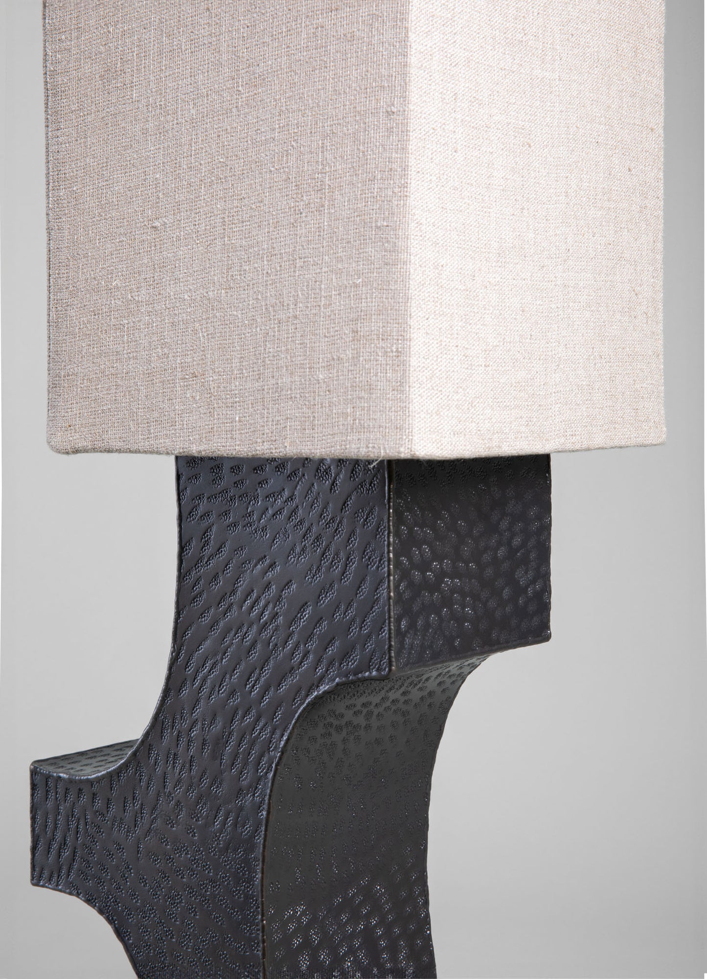 A table lamp with a textured black metal base featuring a hammered pattern, paired with a rectangular cream linen lampshade. The design combines contemporary materials with a minimalist style.