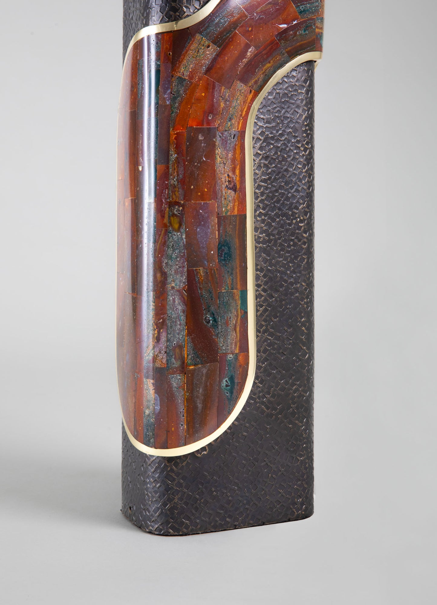 A tall, sculptural artwork featuring a black textured surface with a circular inset of colorful, glazed ceramic tiles in earthy tones, accented with a thin metallic trim. The piece combines modern and handcrafted elements with a focus on geometric shapes and rich, natural colors.