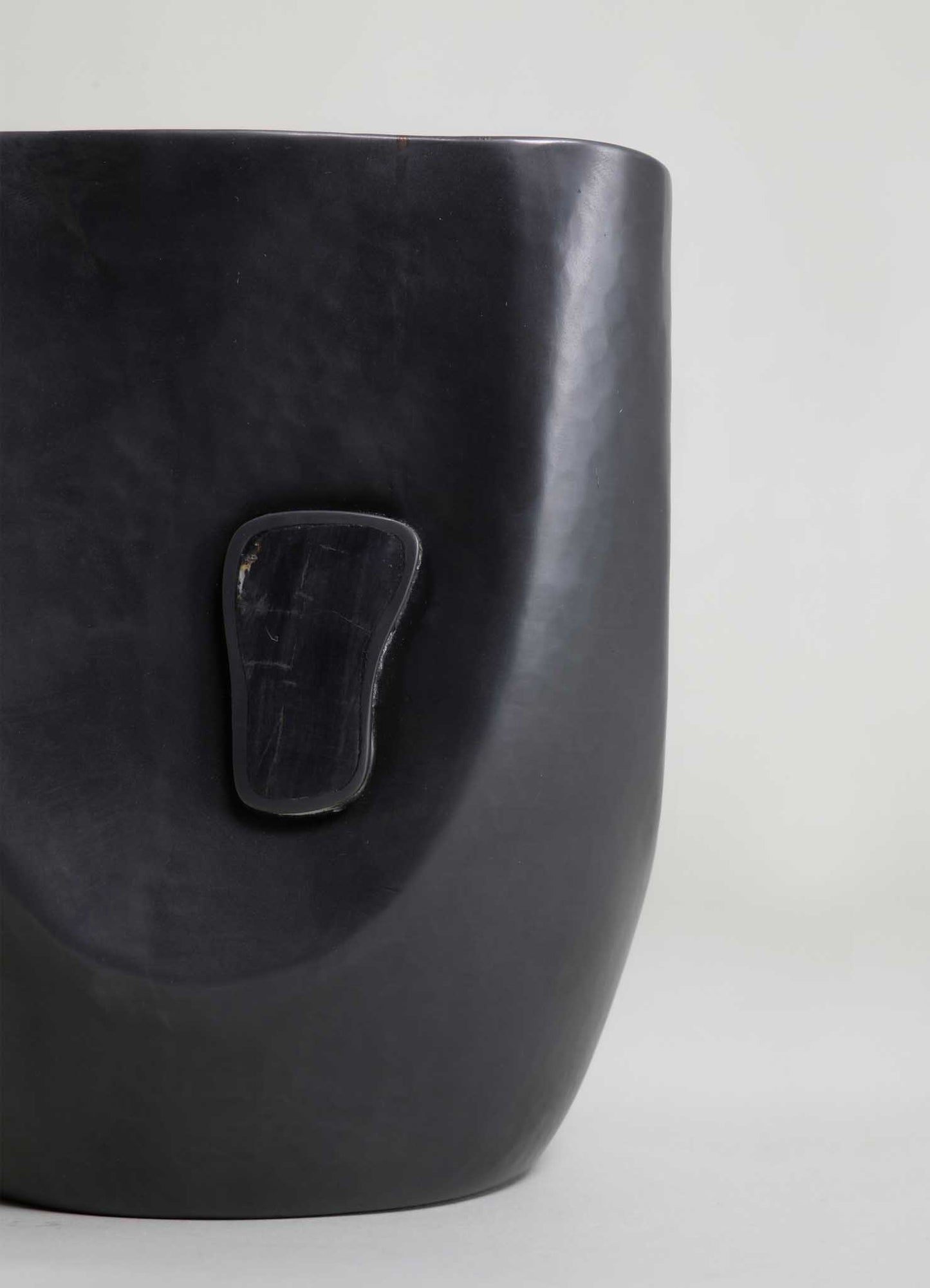A black ceramic or resin planter with a smooth, matte finish and a small, irregularly shaped handle-like detail on its surface. The planter has a contemporary style with a minimalist design.