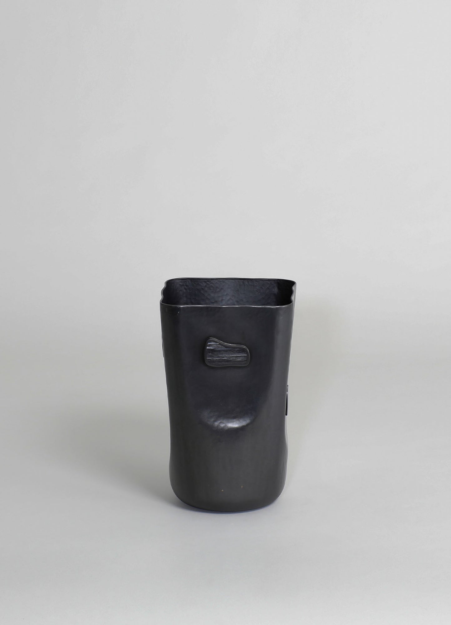 A black ceramic vase with a matte finish, featuring a sculptural, irregular shape and a small rectangular handle on the side. The vase has a modern, minimalist style with subtle textural details.
