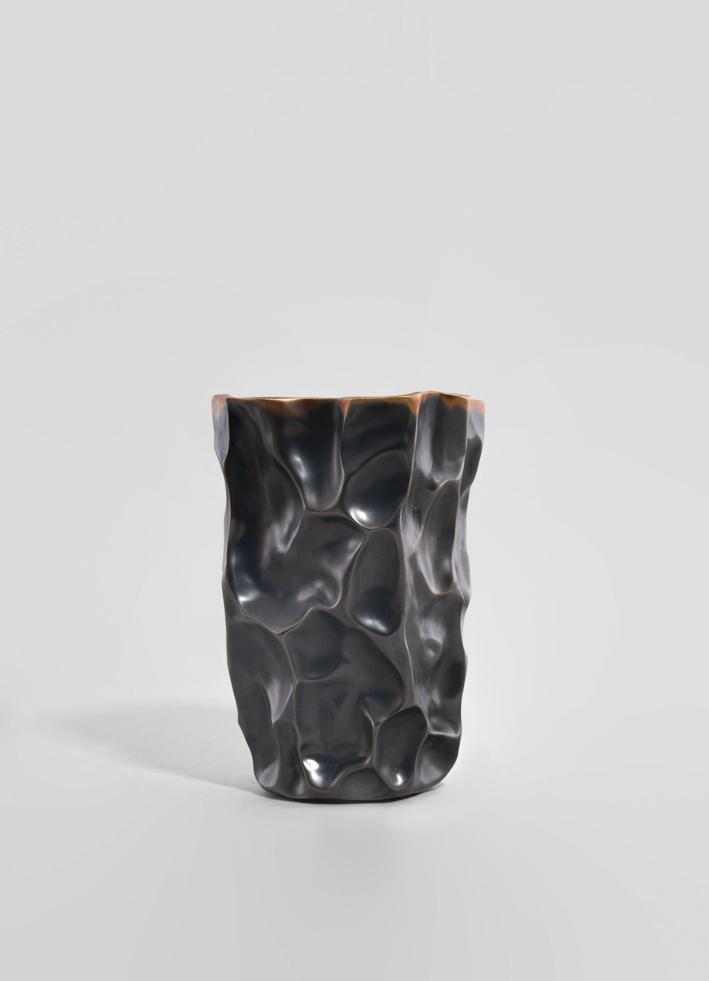A sculptural vase made of dark, polished material with an irregular, organic surface and a glossy finish, set against a plain light gray background.