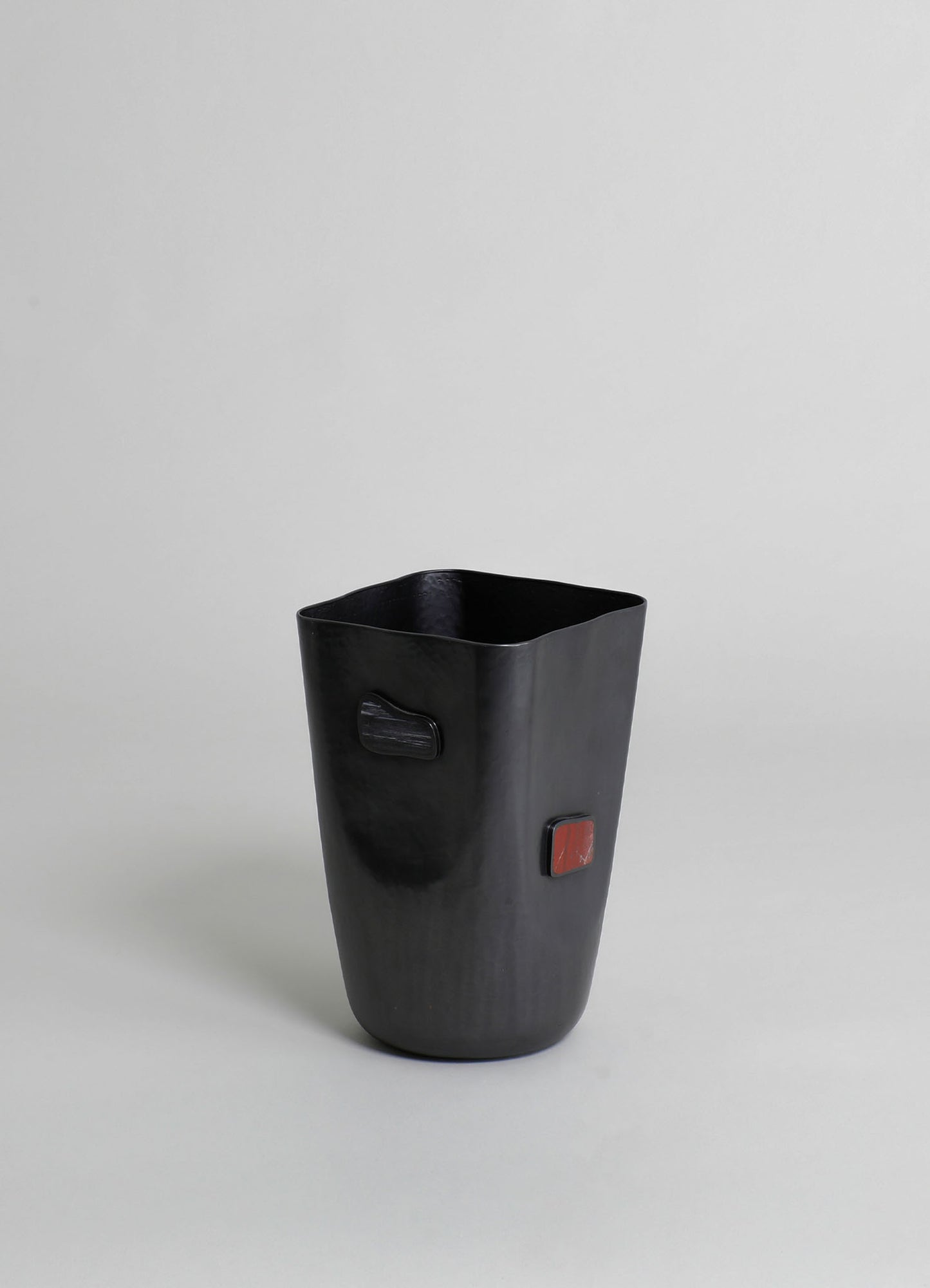 A black ceramic vase with a matte finish, featuring two small rectangular accents in dark gray and reddish-brown on its surface, set against a plain light gray background.