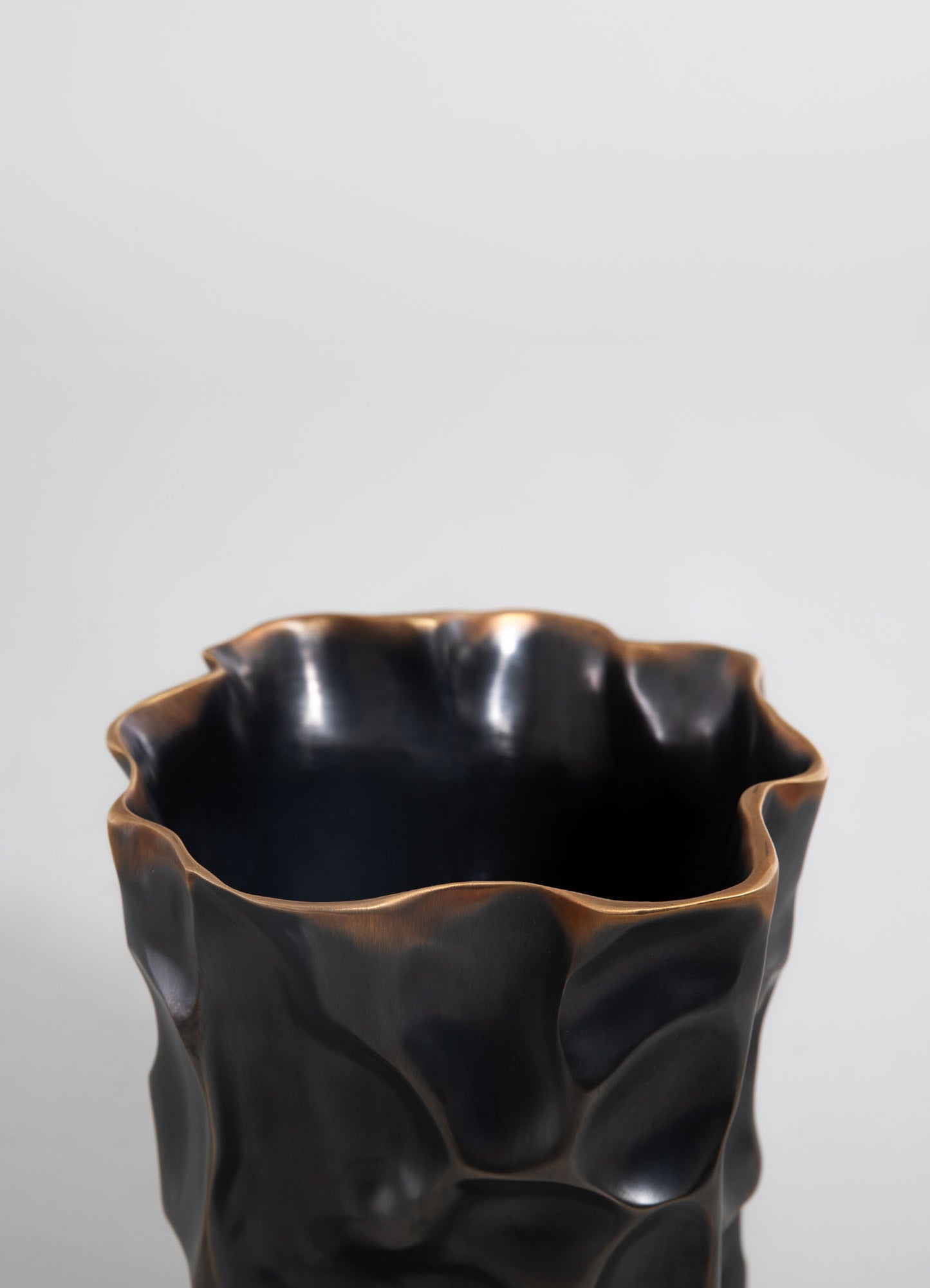 Ceramic vase with a textured, irregular surface in black and bronze tones, featuring a wavy, sculptural form.