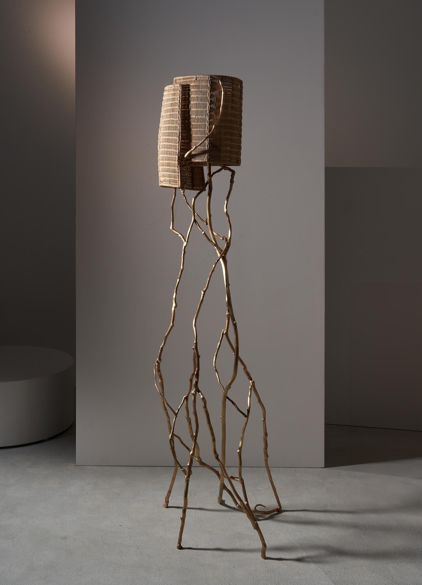 A floor lamp with a woven rattan shade and a sculptural base made of intertwined, natural-colored wooden branches. The design has a contemporary and organic aesthetic, emphasizing natural materials and textures.