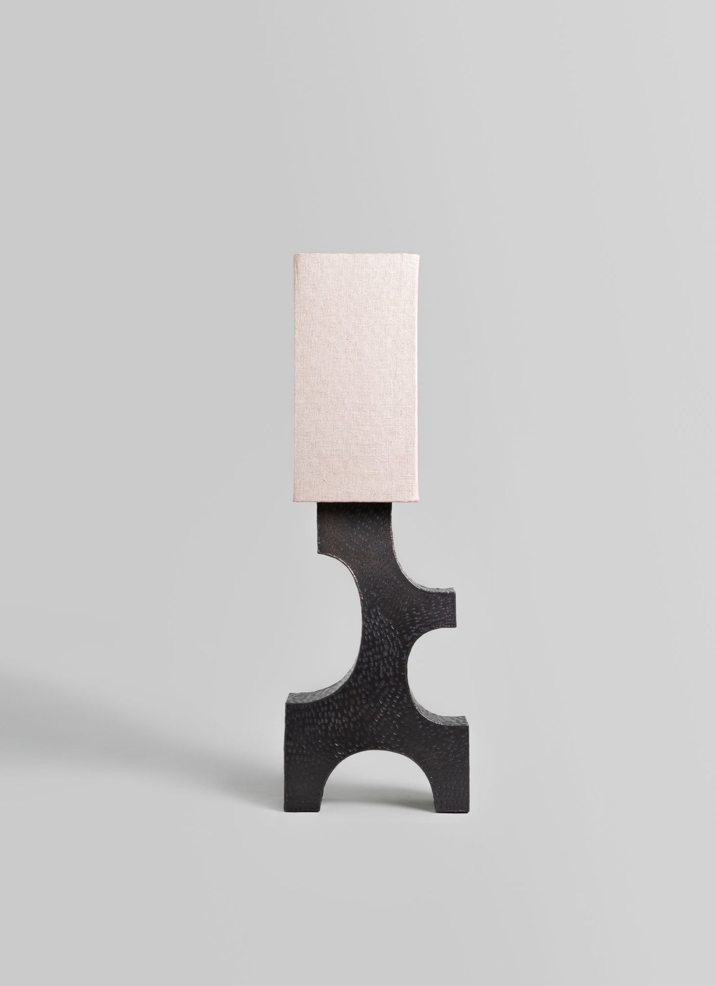 A modern table lamp featuring a sculptural black base with a textured finish and geometric curves, topped with a rectangular fabric lampshade in a light neutral tone.